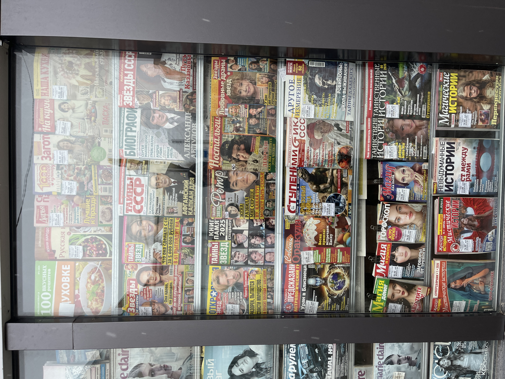 The image shows a kiosk or vending machine filled with various Russian magazines. The magazines cover a range of topics including cooking, biographies of famous people, mysteries, astrology, and general interest stories. The kiosk appears to be located in an urban area, likely in a city in Russia, as evidenced by the Cyrillic text on the magazine covers.