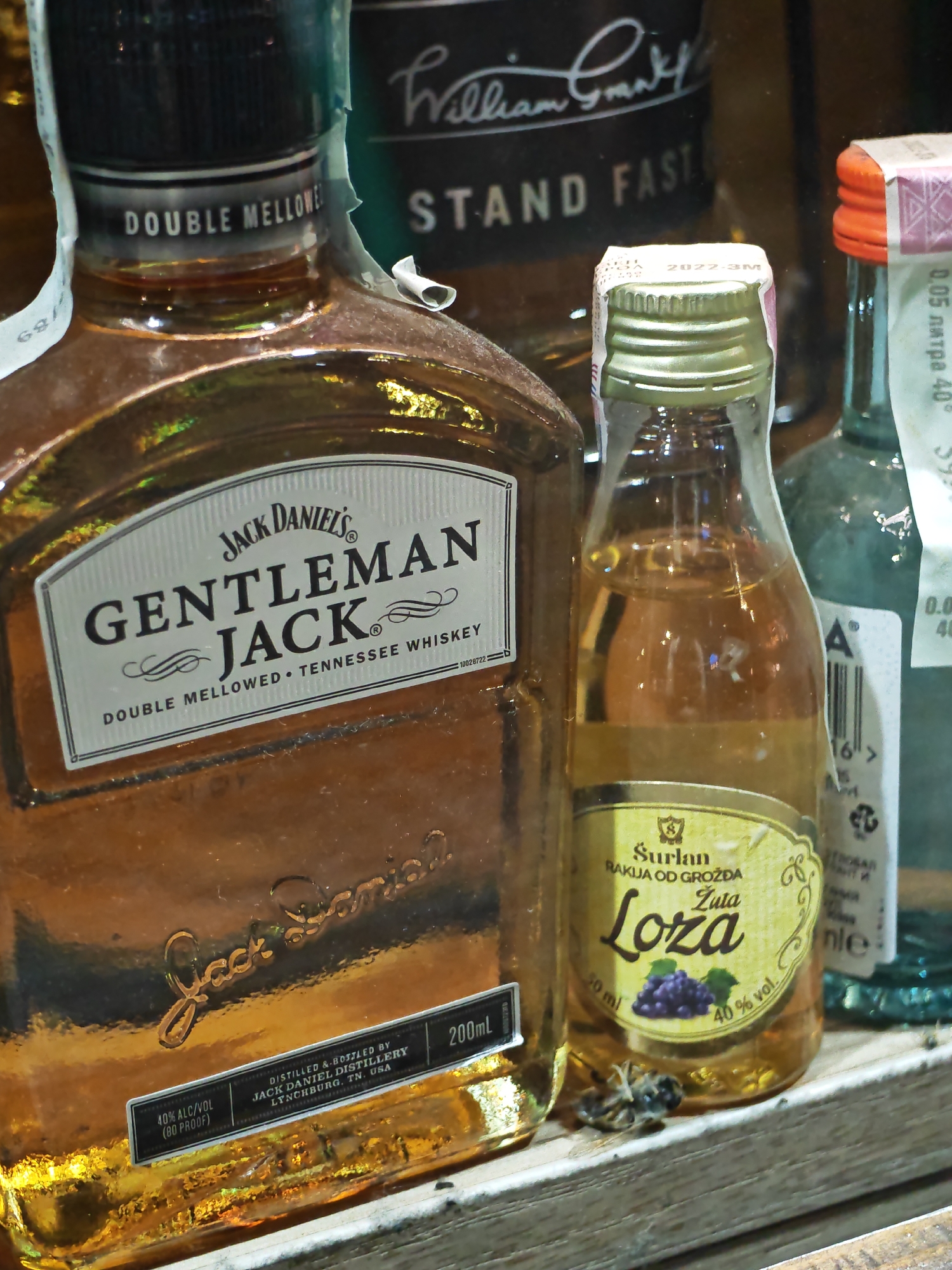 The image shows a close-up of several bottles of alcohol displayed on a wooden surface. In the foreground, a large bottle of Jack Daniel's Gentleman Jack Tennessee Whiskey is prominent. To its right, a small bottle of "Žuta Loza" Rakija od Grožđa is visible. Behind these bottles, parts of other bottles can be seen, including one with a label that appears to read "William Grant & Sons" and another with an orange cap. The lighting suggests an indoor setting, possibly a shelf or display case in a liquor store. A small, dead insect is visible on the wooden surface in front of the "Žuta Loza" bottle.