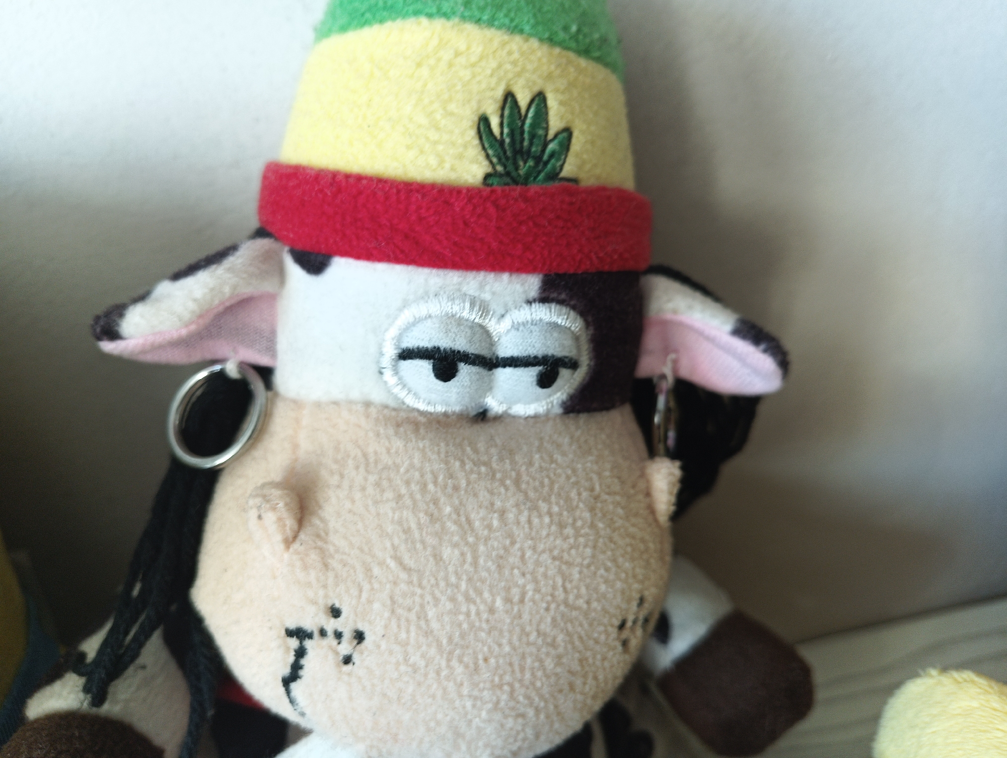 A plush toy cow wearing a hat with red, yellow, and green stripes and a small green leaf design on the yellow band. The cow has black and white patches on its head and pink inner ears. It is wearing a red band around its forehead. The toy has embroidered eyes and nostrils, and what appear to be small stitch marks on its cheeks. It is also wearing a silver hoop earring on its left ear. The cow's body is a light beige fuzzy material, and it has black yarn hair or dreadlocks hanging down. Part of another plush toy, a yellow chick, is visible to the right. The background is a plain, lightly textured, off-white wall. The scene is indoors. There is no indication of the city of Karnobat, Bulgaria.