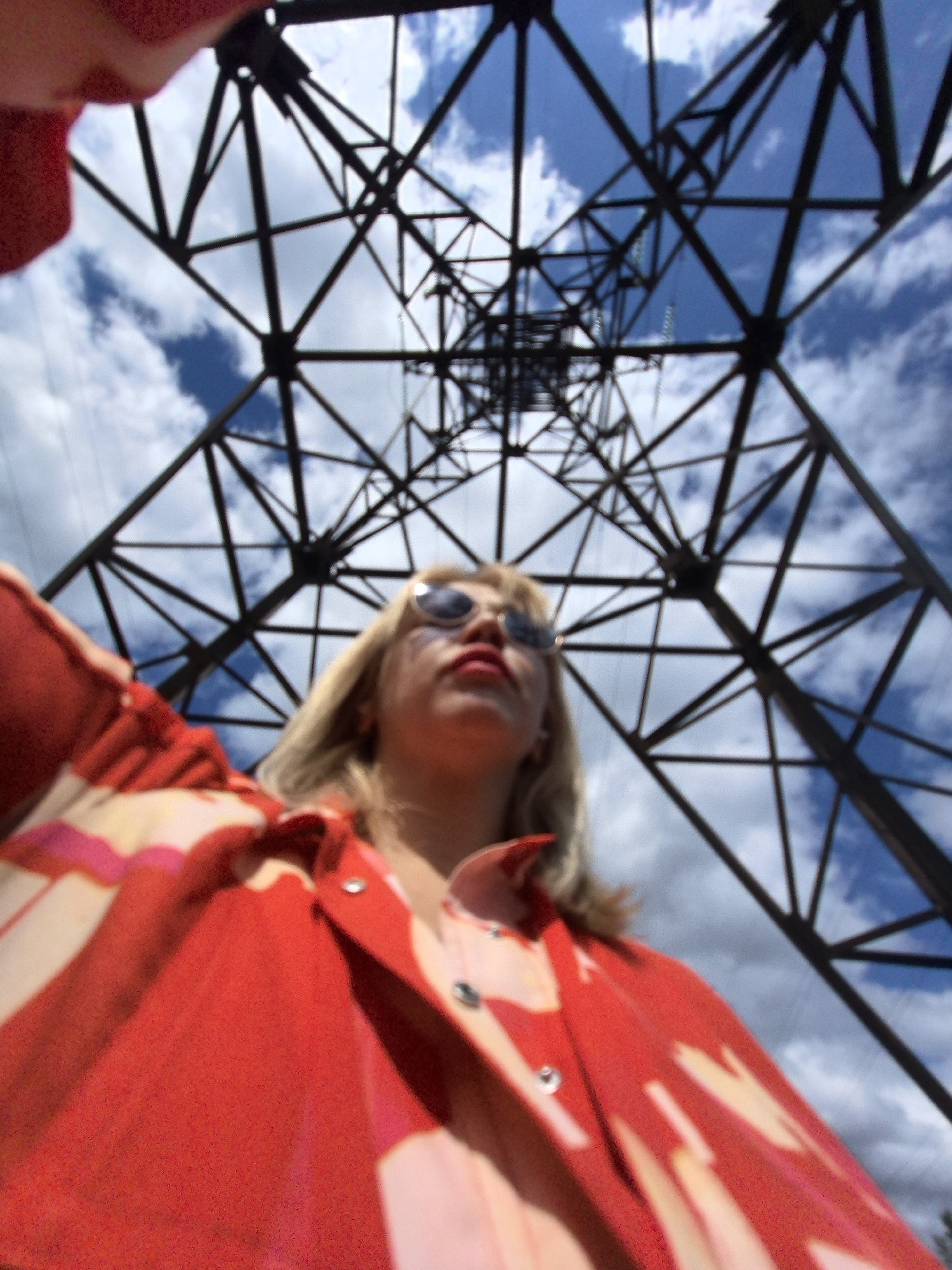 The image shows a person viewed from below, looking up at a large metal tower against a cloudy blue sky. The person is wearing sunglasses and a vibrant red and pink patterned jacket. The camera angle emphasizes the towering structure of the metal framework, which appears to be a power transmission tower. The lighting suggests it is daytime. There are no visible cues in the image to indicate the specific location within Noviy, Russia.