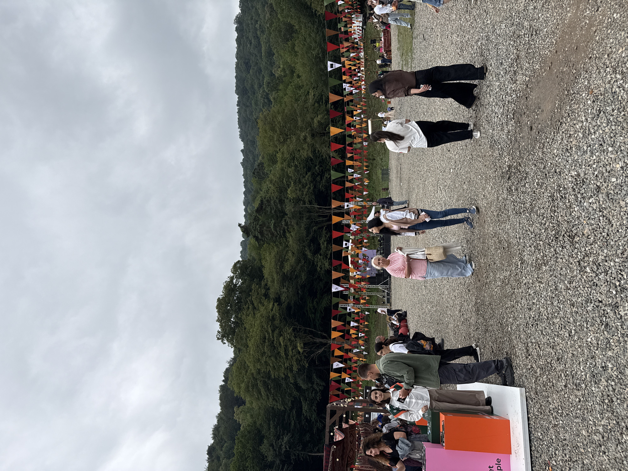 An outdoor festival is taking place under an overcast sky, with a wooded hillside forming the backdrop. Colorful triangular flags are strung across the scene, suggesting a festive atmosphere. Several people are present, some standing and conversing, while others are moving through the area. One group of people is gathered near what appears to be a display or booth, with a pink and orange structure visible. In the background, there are tents and possibly a stage setup. The ground is covered in gravel. The overall scene suggests a community event or gathering in a natural setting.