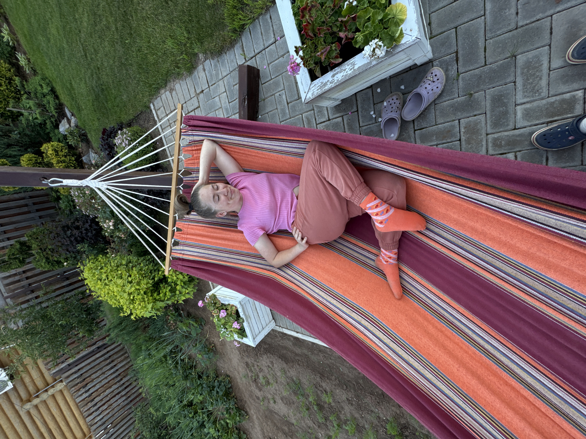 A young woman is relaxing in a striped hammock outdoors in what appears to be a backyard. She is wearing a pink t-shirt and salmon-colored pants with orange socks that have a white pattern. Her head is resting on her arm as she lies in the hammock, which is suspended between two poles. The hammock is patterned with stripes of orange, burgundy, and thin lines of yellow, blue, and white.
The hammock is situated on a paved walkway made of grey rectangular bricks. To the left of the walkway is a garden bed with various plants, including some green leafy plants and some with pink flowers in a white planter. Further left, there is a wooden fence and a grassy slope with more vegetation. To the right of the walkway is a neatly mowed green lawn.
There are two pairs of clogs visible on the walkway, one light purple and another navy blue. One of the purple clogs has a small white flower on it, and there are a few white flower petals scattered on the walkway.
The overall scene suggests a warm, relaxed day in a well-maintained outdoor living space. The mention of "Прикамский, Russia" places this scene geographically, implying it is likely a residential area in that region. The lighting and green foliage suggest it is daytime, likely during spring or summer.