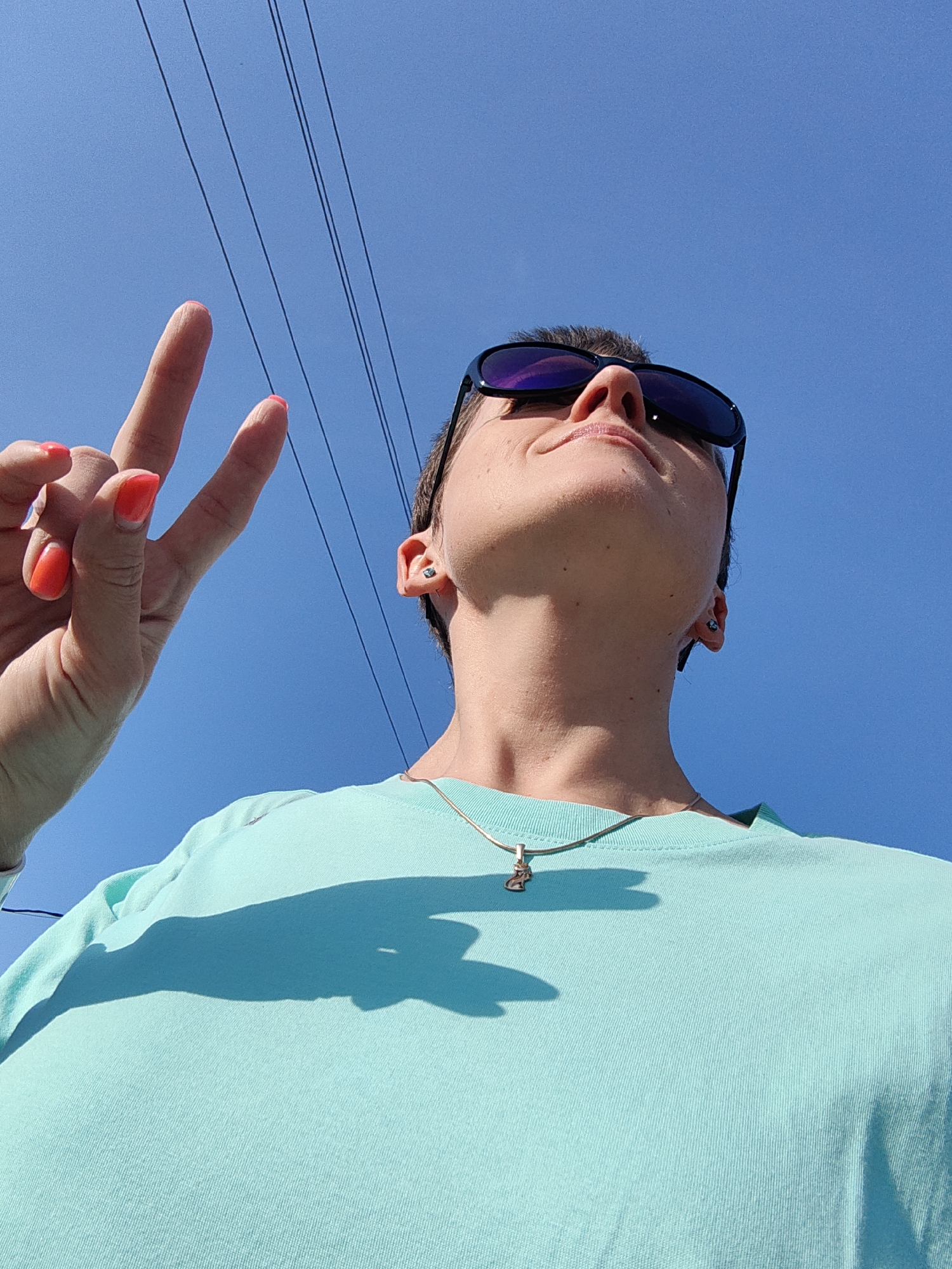 A low-angle, waist-up shot captures a person in a bright turquoise t-shirt and sunglasses against a clear blue sky. The person is making a peace sign with their right hand, their nails painted a vibrant coral. A thin silver chain with a small, ornate pendant rests on their t-shirt. The background features a few dark, thin power lines stretching across the sky. The scene appears to be outdoors, under bright sunlight, casting a distinct shadow of the person onto their t-shirt. The context of Fanipal, Belarus, does not provide any specific visual cues within the image itself.