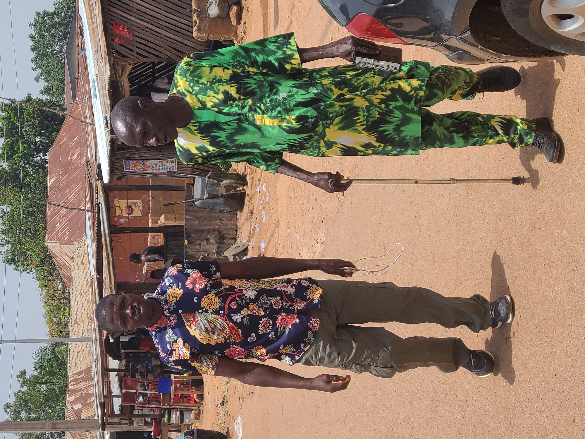 Two men are standing on a dirt street in Jos, Nigeria. The man in the foreground is wearing a blue floral shirt and olive green pants. He is holding white earbuds. Behind him, an older man is wearing a green and yellow patterned suit. He holds a walking stick and a small black electronic device. A car is partially visible on the right. In the background are buildings with corrugated metal roofs.