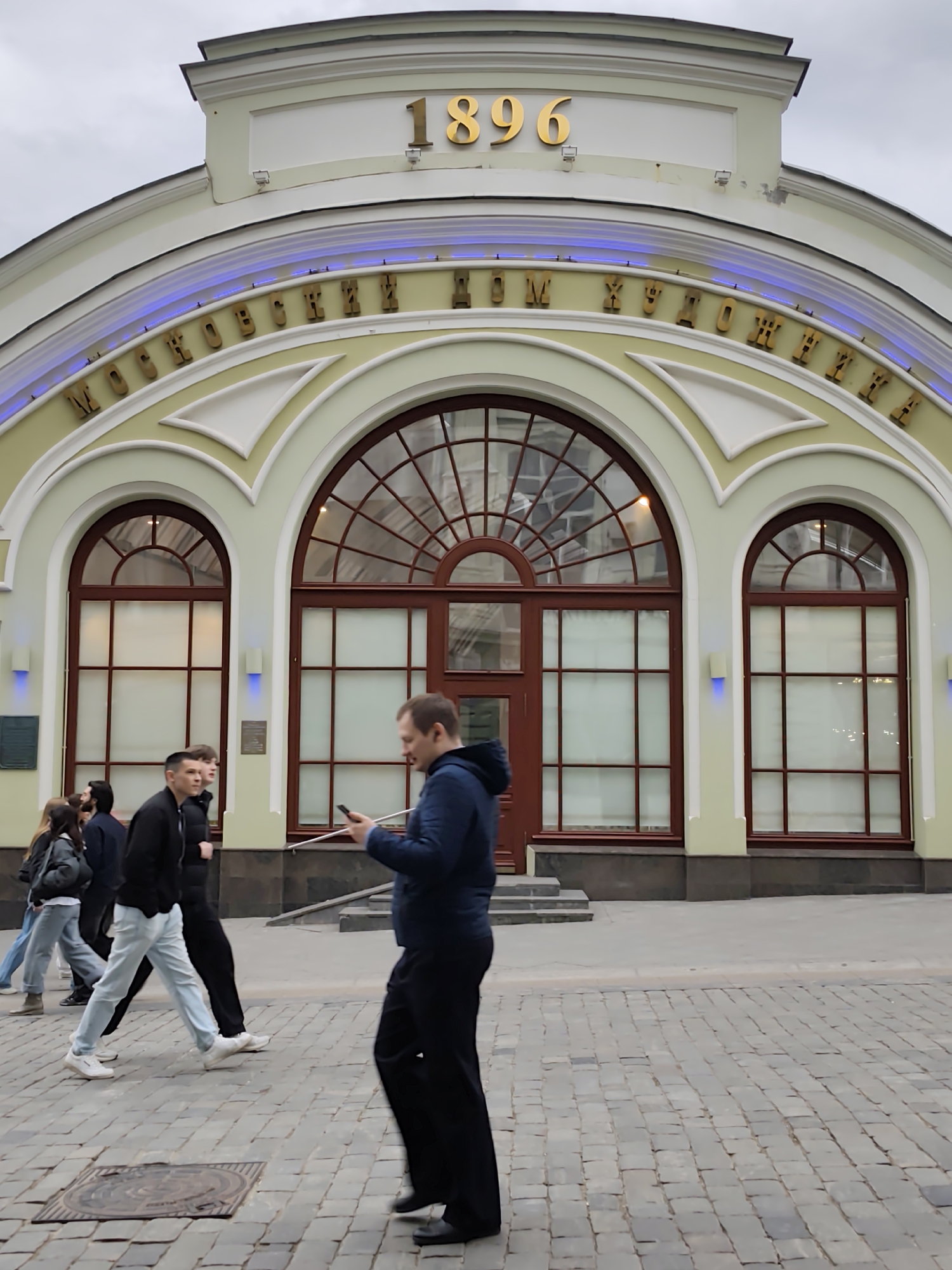 The image shows a street scene in Moscow, Russia, with a building that appears to be a cultural or historical site. The building is light-colored with architectural details including arched windows and the number "1896" in gold lettering above the entrance. The inscription above the arches, in Cyrillic script, translates to "Moscow House of Artists".
In the foreground, several people are walking on a cobblestone street. One man is focused on a handheld device, appearing to be using a smartphone. Other individuals are dressed in casual clothing. The sky appears overcast. The focus is on the building and the people passing by, suggesting a common urban scene.