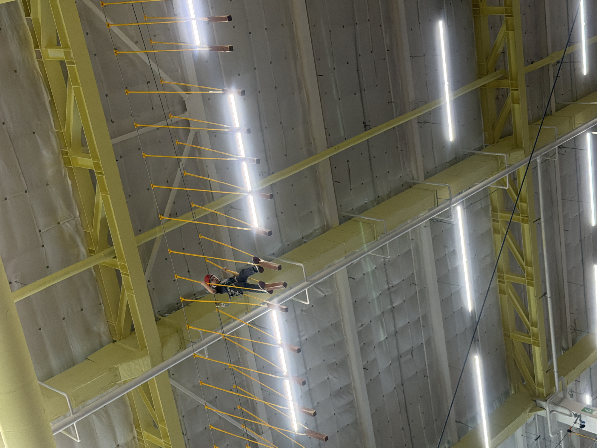 An indoor high ropes course facility in Perm, Russia, is depicted from a low-angle perspective, focusing on an individual traversing an elevated obstacle. A single person, dressed in dark clothing, a dark full-body safety harness, and a red helmet, is observed stepping across a series of dark brown, rectangular wooden blocks suspended by bright yellow ropes. The individual's left foot is firmly placed on one block, while the right foot is lifted, poised to move to the next block. Overhead, a complex industrial ceiling structure is visible, comprising light gray corrugated panels, numerous large yellow-painted metal beams and trusses, and white utility piping. Multiple bright, horizontal LED strip lights are mounted across the ceiling, providing illumination and casting reflections. The ropes course elements are suspended below these structural components.