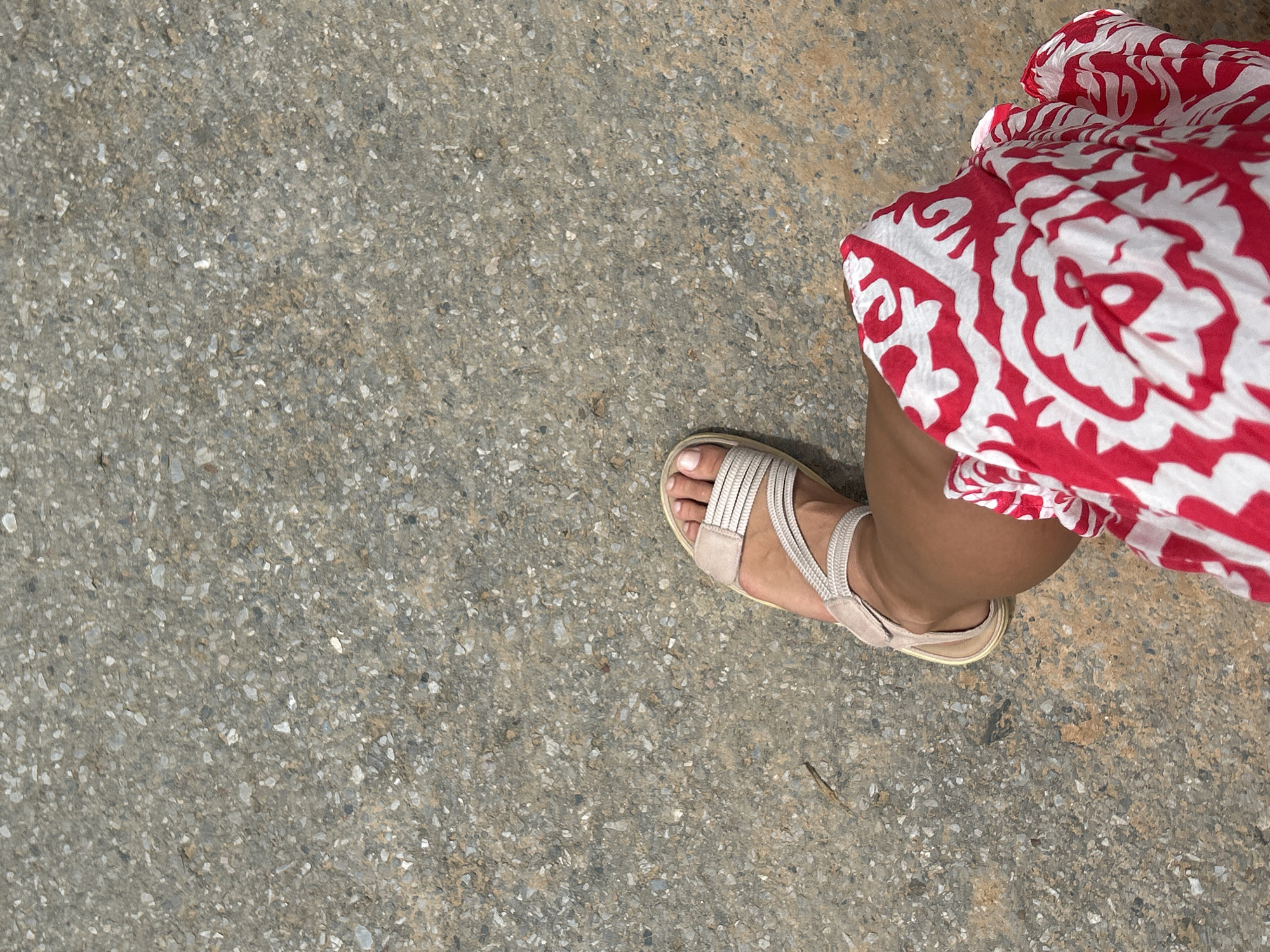 The image is a top-down, close-up shot of a person's right foot and lower leg walking on a textured, grey asphalt surface. The person is wearing a light-colored, strappy sandal. A portion of their leg is visible, covered by a red and white patterned fabric, likely part of a dress or skirt. The pattern is bold and organic, with large white shapes on a red background. The asphalt appears to be a typical outdoor surface, with visible aggregate. The lighting suggests it is daytime, likely sunny, casting no discernible shadows from the foot or sandal. The scene is a simple, everyday moment of walking. There is no visible text in the image. The location context is Kathu, Thailand, which implies a warm, possibly tropical environment.
