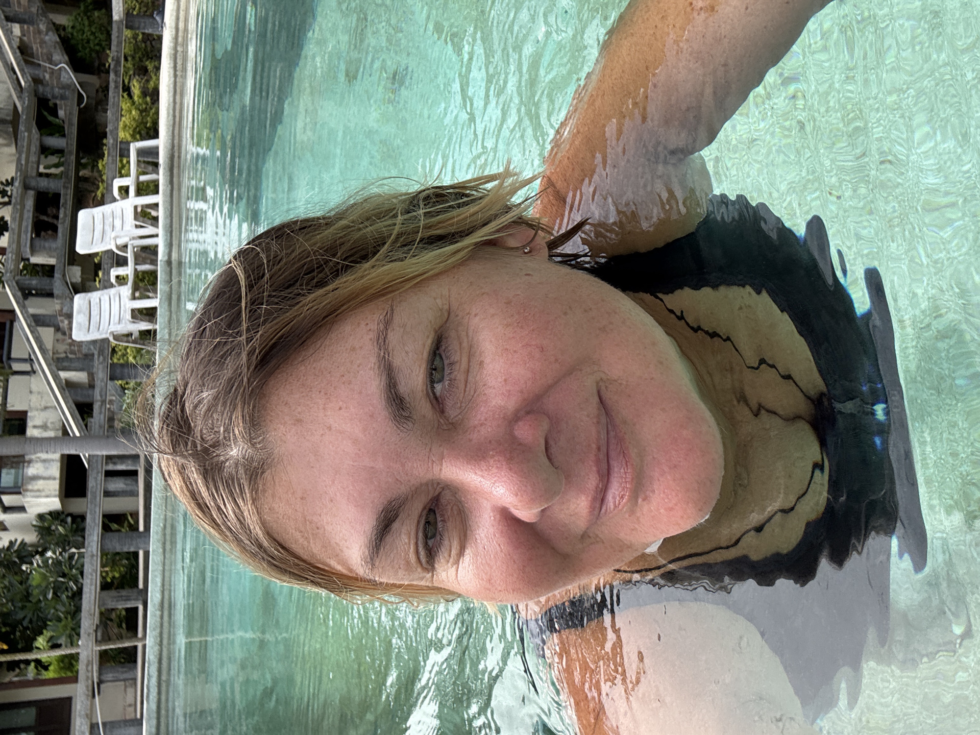 The image shows a woman in a swimming pool. She is facing the camera with her face partially submerged. The pool is turquoise and the water is rippling. In the background are white lounge chairs and a building. The scene is likely in บ้านบางรักษ์, Thailand, due to the prompt's requirement.