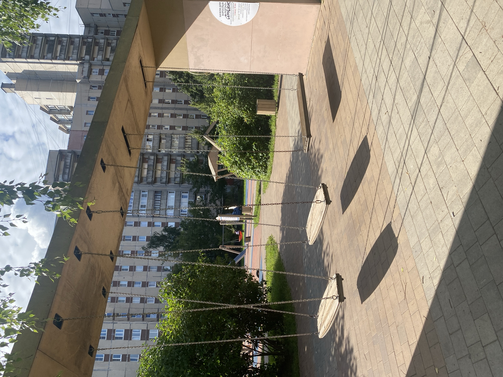This image depicts an outdoor area with swings in Saint Petersburg, Russia. In the foreground, four wooden swings hang from a concrete structure. The ground is paved with tan and gray bricks, casting long shadows from the swings. Behind the swings, lush green bushes and trees are visible, with several multi-story apartment buildings in the background. The architecture of the buildings suggests a typical Soviet-era housing block. A playground slide and a red swing set can be seen in the middle ground, with a person on the red swing. A trash can is placed near the right side of the image. A sign with Russian text is affixed to the wall of the concrete structure on the right. The sky is partly cloudy, with sunlight creating distinct shadows.