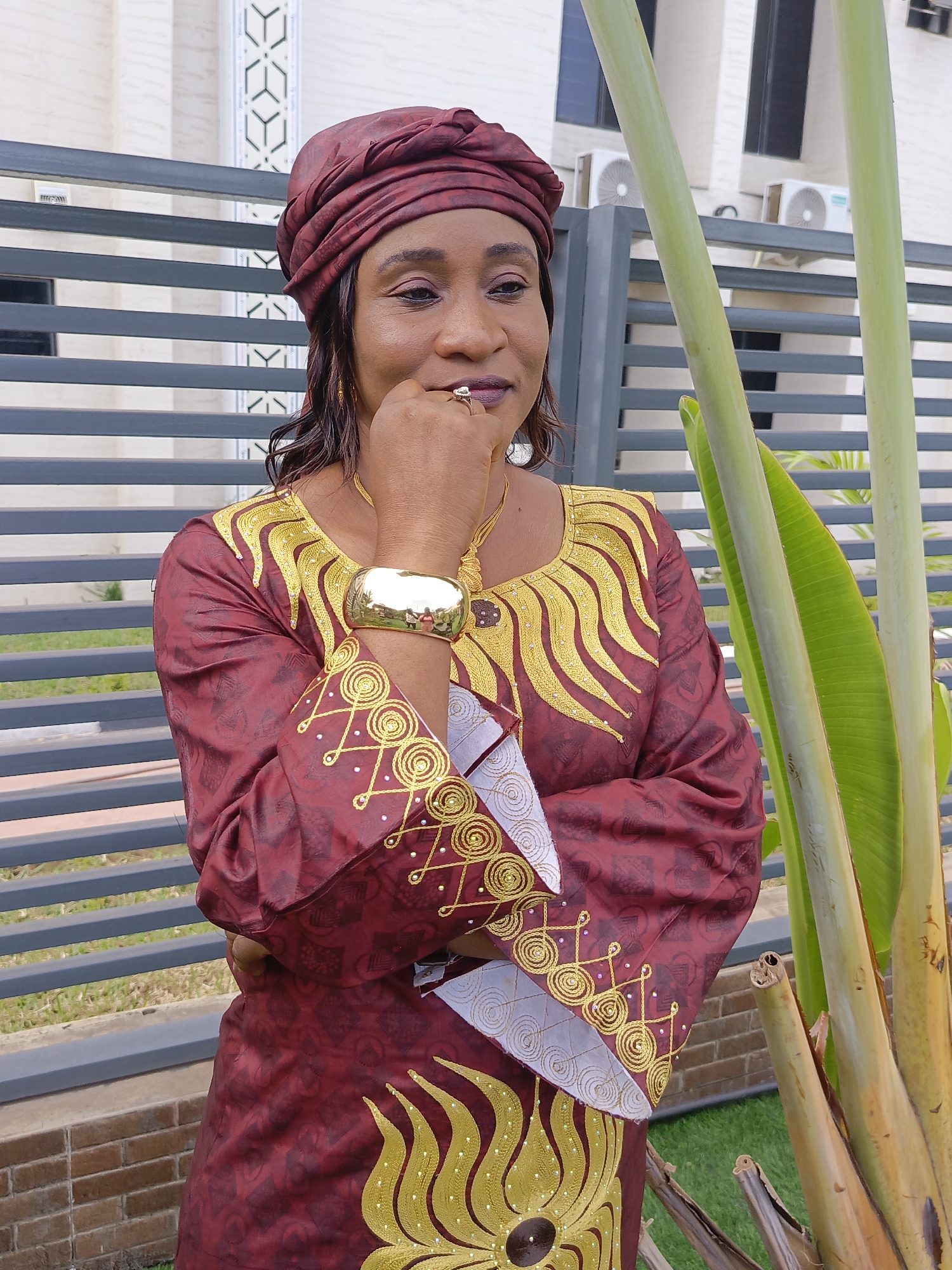 A portrait-style image captures a woman in traditional attire, standing outdoors in Akwanga, Nigeria. She is positioned slightly off-center to the right, looking towards the right with a contemplative expression, her right hand gently touching her chin.
The woman is wearing a striking maroon garment, possibly a bubu or a similar traditional dress, richly embellished with intricate gold embroidery. The chest area features bold, wave-like gold patterns, while the sleeves showcase circular and geometric designs accented with small white beads. A matching maroon headwrap adorns her head. Her accessories include a large, shiny gold cuff bracelet on her left wrist, a gold-colored necklace, gold earrings, and a ring on her right hand.
In the foreground on the right, large, thick green stems and leaves of a tropical plant, possibly a banana plant or a traveler's palm, partially frame the scene. The base of this plant shows dried, brown remnants. Behind the woman, a modern dark grey slatted fence or gate extends across the midground. Beyond the fence, a building with light-colored walls and dark-framed windows is visible. Two white air conditioning units are mounted on the building's exterior. To the far left, another part of a building with a distinctive white geometric pattern adds to the background. Green grass and a brick wall are visible at the lower left.
The scene is brightly lit, suggesting daytime. The overall ambiance is calm and thoughtful. Faint text "31A" is visible on one of the plant stems, and "SIEMENS" can be seen on one of the air conditioning units.
