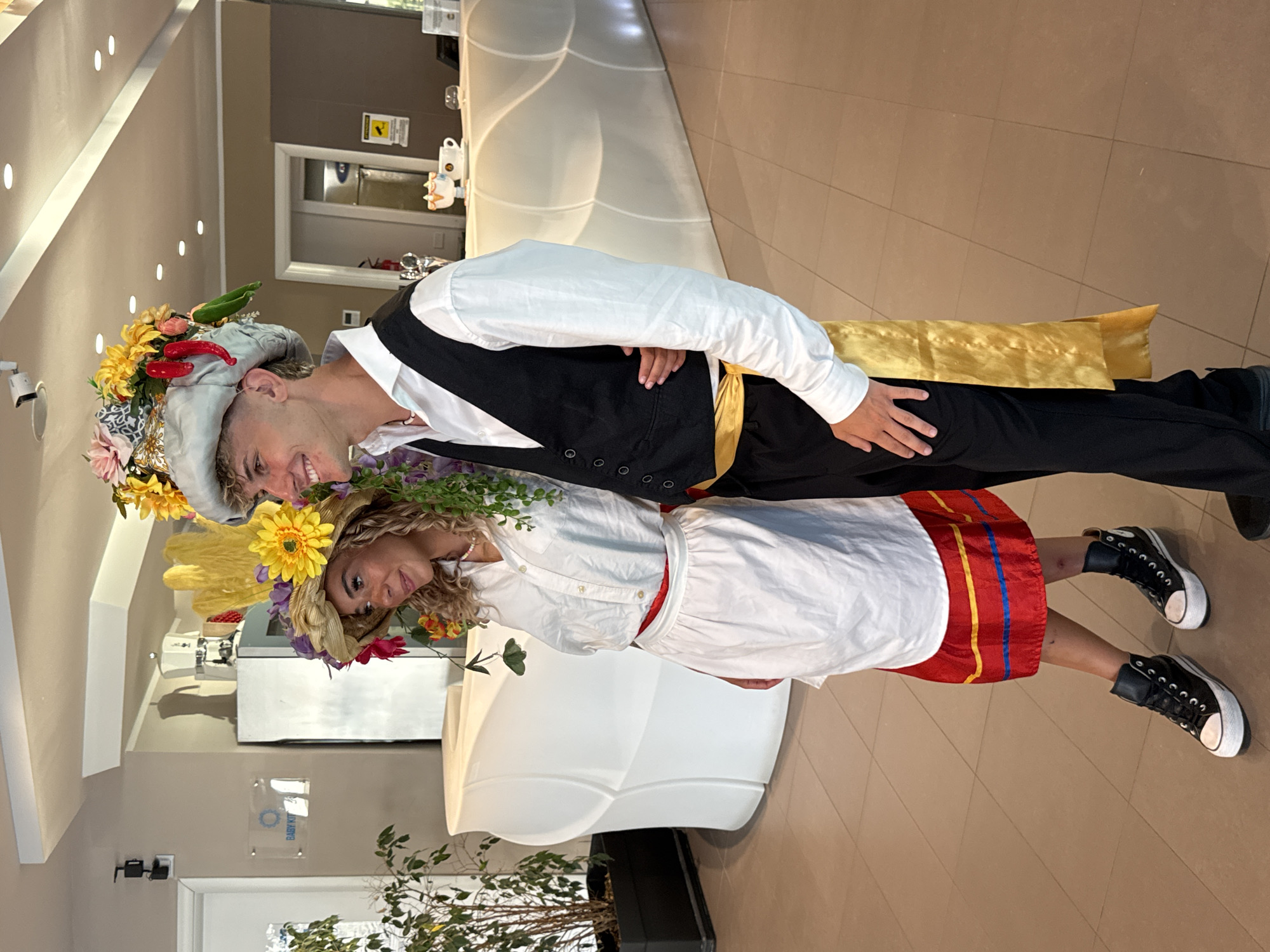 Two young adults, a male and a female, are posing for a photograph indoors. They are both wearing elaborate, decorative hats. The female, on the left, has on a straw hat adorned with yellow and pink flowers and green foliage. She is wearing a white shirt, a white apron over a red skirt with yellow and blue trim, and black high-top sneakers. The male, on the right, is wearing a grey cap-like hat with a colorful assortment of flowers and red chili peppers attached. He has on a white dress shirt, a black vest, black pants, and a golden sash tied around his waist. They are standing in front of a white, modern reception desk. The background shows a wall with a framed sign that says "BABY KIT" and a potted plant. The flooring is tiled. The location is the interior of a hotel or reception area in Isola delle Femmine, Italy.