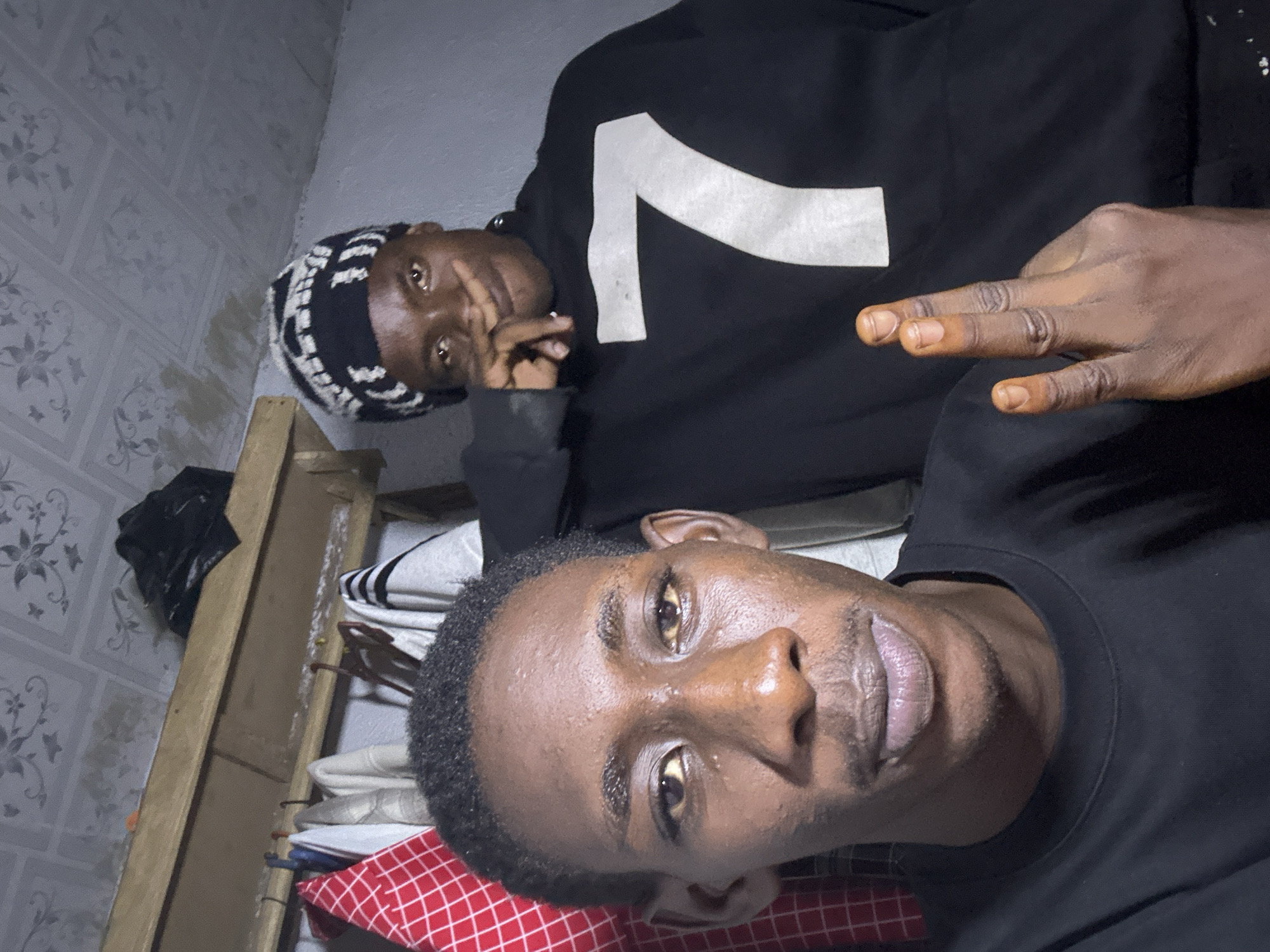 In a dimly lit room, possibly a closet or storage area, two young individuals are captured in a selfie. The person in the foreground, positioned on the left and facing the camera directly, is a young male with dark skin, short black hair, and a faint mustache. He is wearing a black t-shirt. His gaze is directed towards the camera, with his face filling the frame. Behind him, slightly to the right and further back, stands another young individual wearing a black beanie with a white patterned design and a black sweatshirt featuring a large white number "7" on the chest. This individual is partially obscured, with their face turned towards the camera and their hand raised, touching their nose or mouth area.
The background reveals a cluttered environment. Above the individuals, a wooden shelf is visible, holding a black plastic bag and various items of clothing hung behind them. The ceiling appears to be covered with a patterned material. The overall impression is one of an informal, personal space. There are no distinct landmarks or environmental cues to suggest a specific location within Durumi, Nigeria, beyond the implication of an indoor setting.