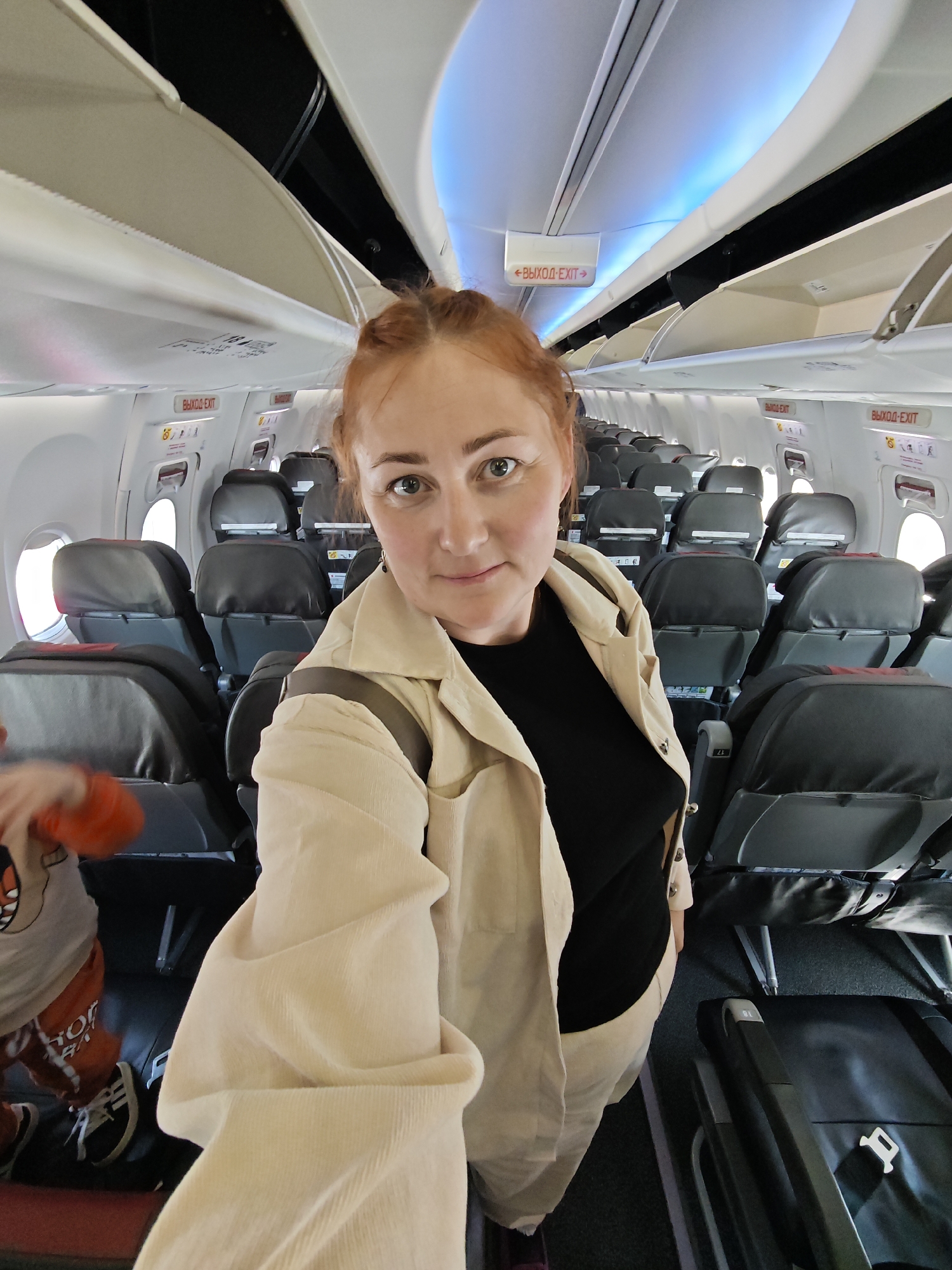 The image captures the interior of a passenger aircraft cabin, presenting a selfie taken by a woman. The environment is characterized by rows of dark grey passenger seats with subtle red accents, a dark grey carpeted aisle, and circular windows visible on the left side. Overhead bins, some open and some closed, line the ceiling, which features both white cabin lighting and a strip of blue accent lighting. Prominently displayed throughout the cabin are red "ВЫХОД-EXIT" signs, containing both Russian (ВЫХОД) and English text, indicating a Russian operational context for the aircraft. This setting is consistent with an aircraft operating in Saint Petersburg, Russia.
In the foreground, a woman with reddish-brown hair, appearing to be in her 30s or 40s, is taking the photograph. She wears a light beige corduroy jacket over a black top and light-colored pants. Her eyes are light-colored, and she has a small earring in her left ear, with a neutral to slightly smiling expression as she looks directly at the camera. Partially visible in the lower-left corner, blurred from motion, is a young child. The child wears an orange long-sleeved shirt with an indistinct graphic, orange shorts with white lettering, and black and white sneakers. The child appears to be standing or moving in the aisle near the woman. A black hardshell suitcase rests on the aisle floor in the bottom right quadrant of the image.