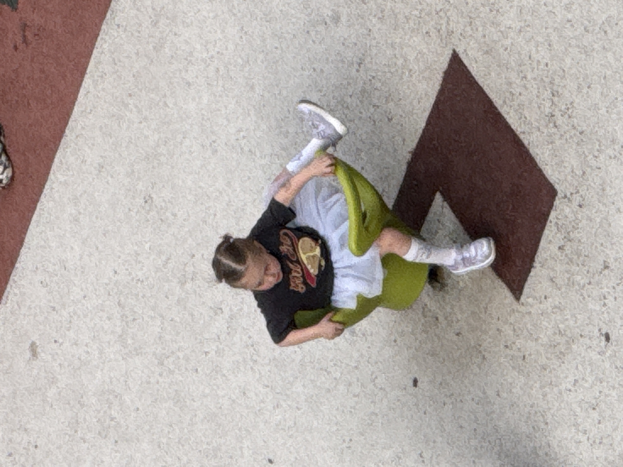 An overhead shot shows a young person sitting on a bright green chair on a speckled, light-colored ground, which appears to be pavement or concrete. The person is wearing a black t-shirt with a graphic design, light-colored shorts or skirt, white socks, and grey sneakers. Their hair is pulled back in a bun. They are holding onto the chair, with one leg bent and the other extended.
To the left of the person, a reddish-brown surface, possibly a wall or a structure, occupies a portion of the frame. To the right and behind the chair, a dark brown diamond-shaped pattern is visible on the ground. The lighting suggests it is daytime, and there are no immediate indicators of the weather conditions. The overall scene appears to be an outdoor public space, possibly a playground or a park in Moscow, Russia. No discernible text is visible in the image.
