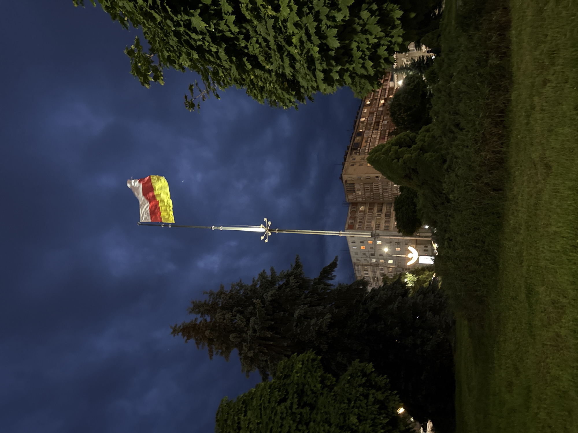 This media file shows a nighttime view of a city scene with a flag in the foreground. The flag, which appears to be composed of white, red, and yellow stripes, is fluttering on a pole against a dark, cloudy sky. Behind the flag, a tall building with illuminated windows is visible, suggesting an urban setting. Trees and manicured green grass flank the building and the flagpole, creating a park-like atmosphere. A street lamp with a unique star-like fixture is also present, casting light on the surroundings. The overall mood is serene and somewhat mysterious, amplified by the low light conditions and the vast expanse of the night sky. The image has been rotated, with the flag and building appearing sideways. The setting appears to be Vladikavkaz, Russia.