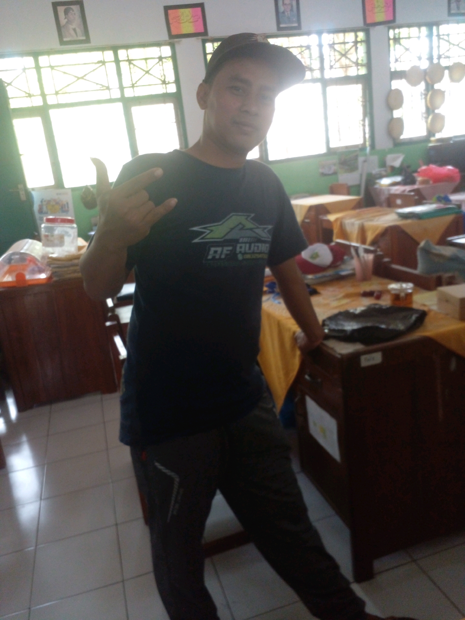 A young adult male, wearing a dark t-shirt and dark pants, stands in a classroom setting in Banyuputih, Indonesia. The man is facing forward, with his left hand raised in a gesture. In the background, there are desks with yellow tablecloths, windows, and various items on the desks and walls, including framed pictures and containers. The lighting suggests it is daytime, with sunlight streaming through the windows.