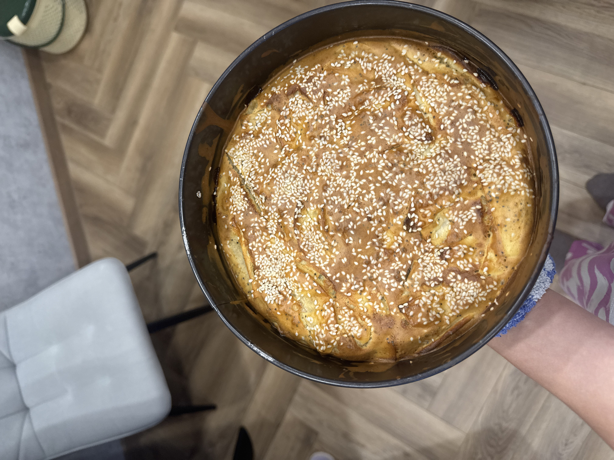 An overhead shot captures a freshly baked, round item in a dark-colored pan, held by a person's hand and arm. The baked good has a golden-brown, slightly irregular surface, generously sprinkled with white sesame seeds. Some darker specks, possibly poppy seeds or herbs, are also visible within the crust. The item appears to be a savory pie, quiche, or a type of cake, filling the dark, round baking pan.
Only a portion of the person holding the pan is visible, specifically their right arm and hand, wearing a light-colored, possibly patterned, oven mitt or sleeve near the wrist. The pan is held over a light-colored wooden floor, which features a distinct herringbone pattern, suggesting an indoor setting like a kitchen or dining area.
In the background, to the left, a light gray upholstered chair with a tufted back and dark legs is partially visible. Further in the top left corner, part of a green object, possibly a hat or a woven basket with a lighter stripe, can be seen. The lighting suggests an indoor environment, but the time of day is not discernible. No text is visible in the image.