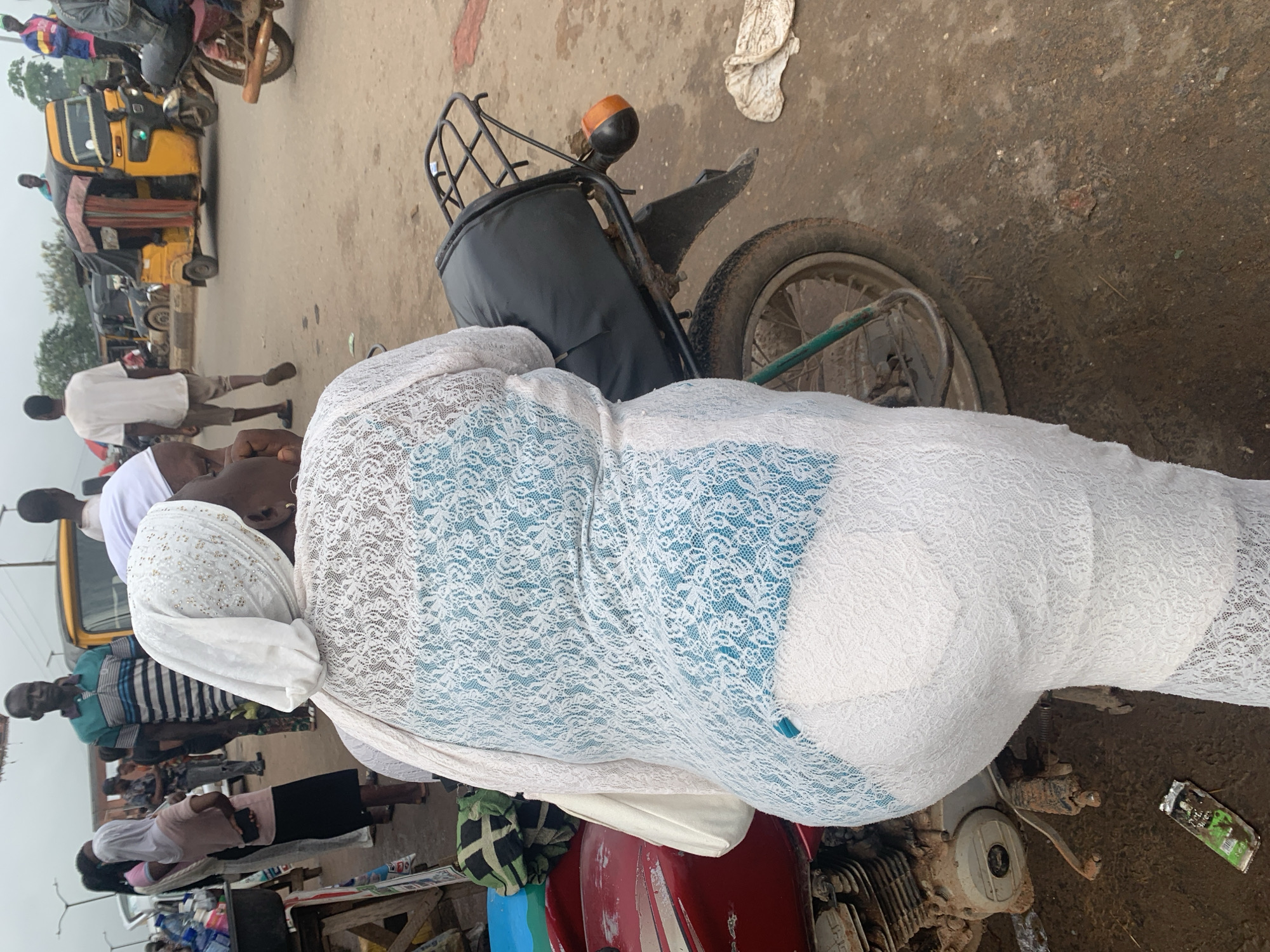 In Shagamu, Nigeria, a medium shot captures a woman from behind, seated on a red motorcycle with a black passenger seat and a black luggage rack. She is wearing a white lace overlay over a light blue garment, with a white headscarf. To the left of the motorcycle, a portion of a red object and a green and black patterned fabric are visible. In the background, several people are walking on a dirt road, with a yellow motorized tricycle (Keke Napep) and other vehicles on the street. The overall environment appears to be a busy marketplace or transportation hub.