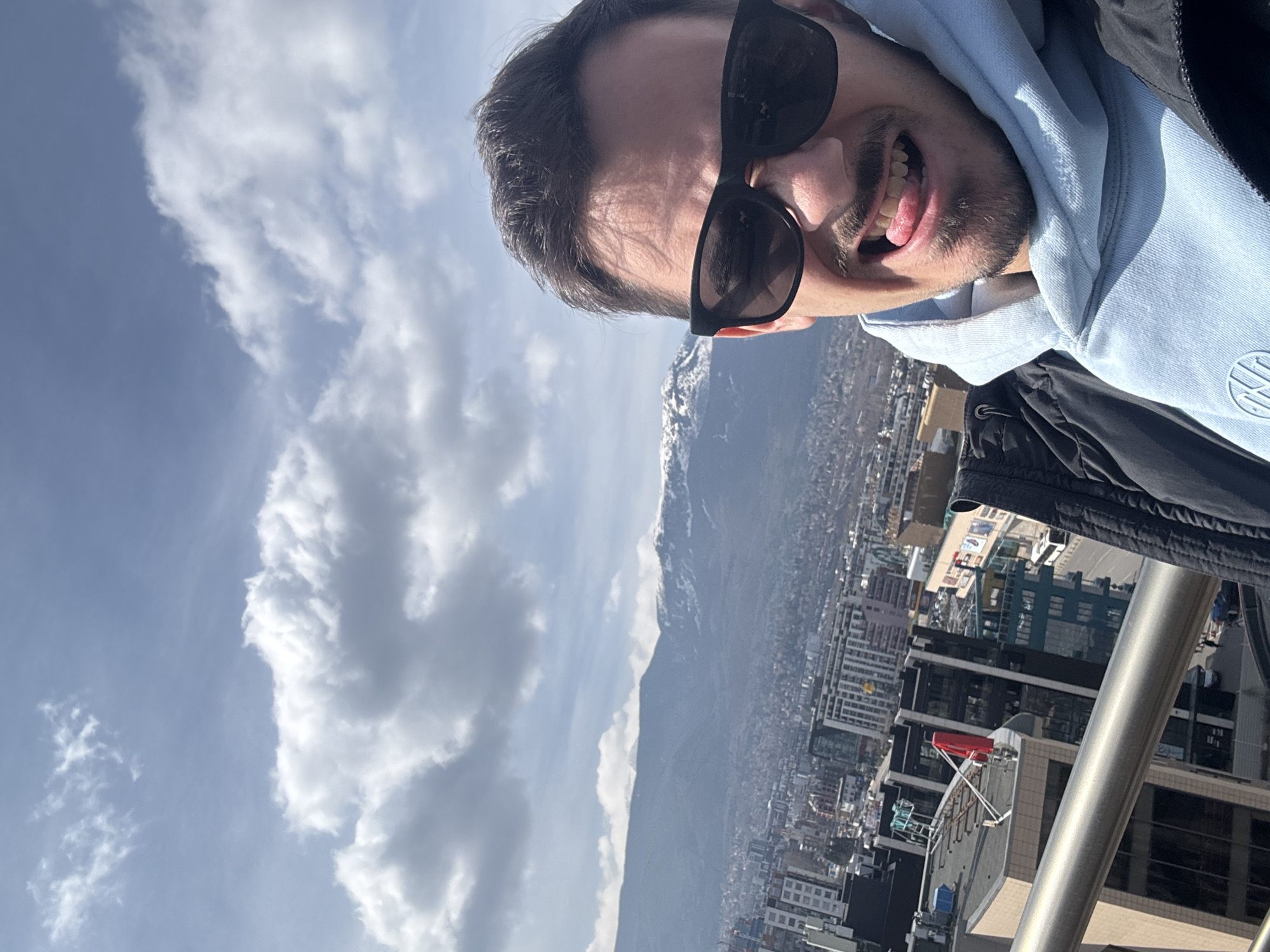A male individual is captured in a close-up selfie from a high vantage point overlooking the city of Sofia, Bulgaria, with Vitosha Mountain in the background. The man, with dark hair, a mustache, and a short beard, wears dark rectangular sunglasses, a light blue hooded sweatshirt featuring a circular logo, and a dark jacket. He is smiling with his tongue slightly extended. A silver metal railing occupies the lower left foreground. Below the railing, the urban landscape of Sofia is visible, featuring numerous buildings of varying heights, including modern glass-fronted structures and rooftops. One prominent rooftop displays a distinct red, angular structure, possibly an antenna or sign. A street with vehicles, including a white bus, is discernible. In the background, the Vitosha Mountain range rises, its higher peaks partially covered with snow. The sky is partly cloudy with significant white cumulus cloud formations against a blue background, indicating daytime.