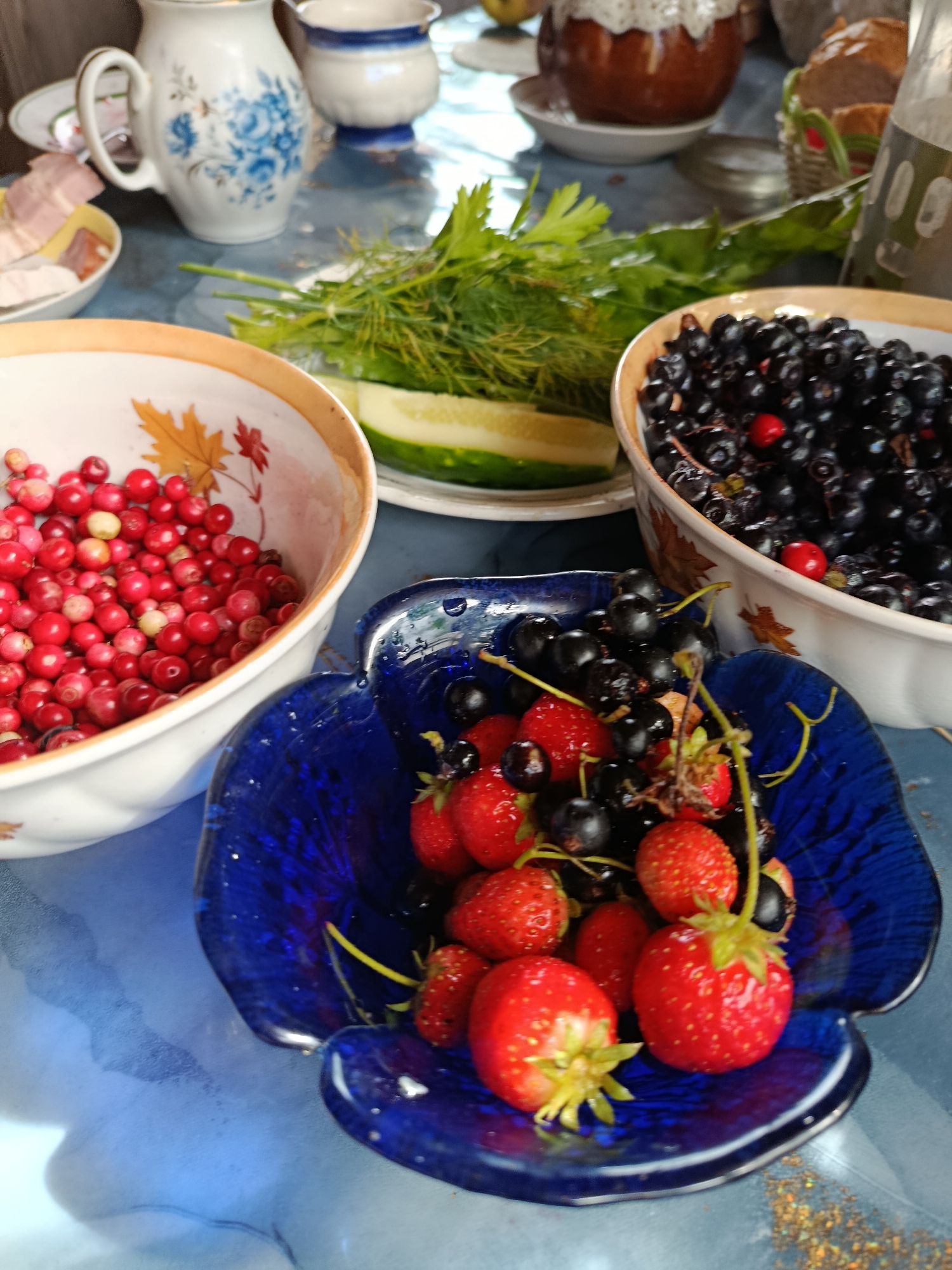 A close-up, eye-level shot showcases a table laden with berries and other food items, suggesting a meal or gathering. In the foreground, a striking blue, heart-shaped bowl overflows with a vibrant assortment of fresh strawberries and dark cherries, some still attached to their stems. To the left, a white bowl with a decorative rim, featuring autumnal leaf motifs, holds a generous portion of small, red berries, likely lingonberries or cranberries. Behind these bowls, a larger white bowl is filled with a dark mass of berries, appearing to be blackcurrants. A serving of fresh dill and sliced cucumber rests on a plate between the bowls, adding a touch of green to the scene. In the background, a white pitcher with blue floral detailing stands prominently, accompanied by other indistinct kitchenware and a basket of bread. The overall setting appears to be an indoor table, possibly in a domestic environment, with a patterned blue tablecloth as the base. The lighting suggests natural daylight filtering in. The image captures a moment of abundance and the simple pleasure of fresh, seasonal produce.