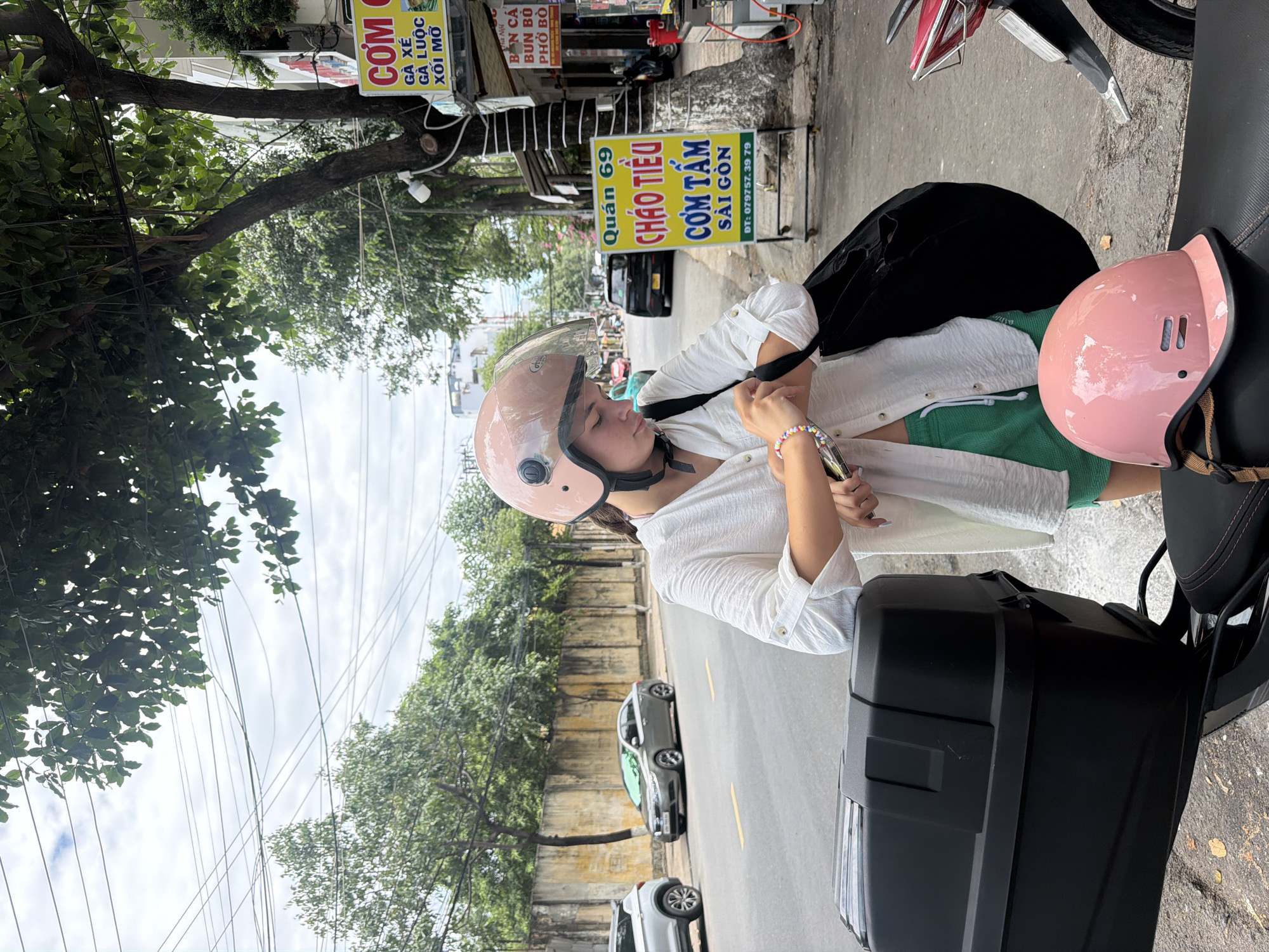 A young woman wearing a pink helmet and a white shirt stands on a sidewalk in Nha Trang, Vietnam, adjusting her watch. She has green shorts visible beneath her shirt and is carrying a black bag. In the foreground, a black motorcycle with a storage box is parked, and a pink helmet rests on its seat. Behind her, a yellow sign displays Vietnamese text, likely indicating a food establishment. The street is paved, with parked cars and trees lining the road. The sky is overcast.