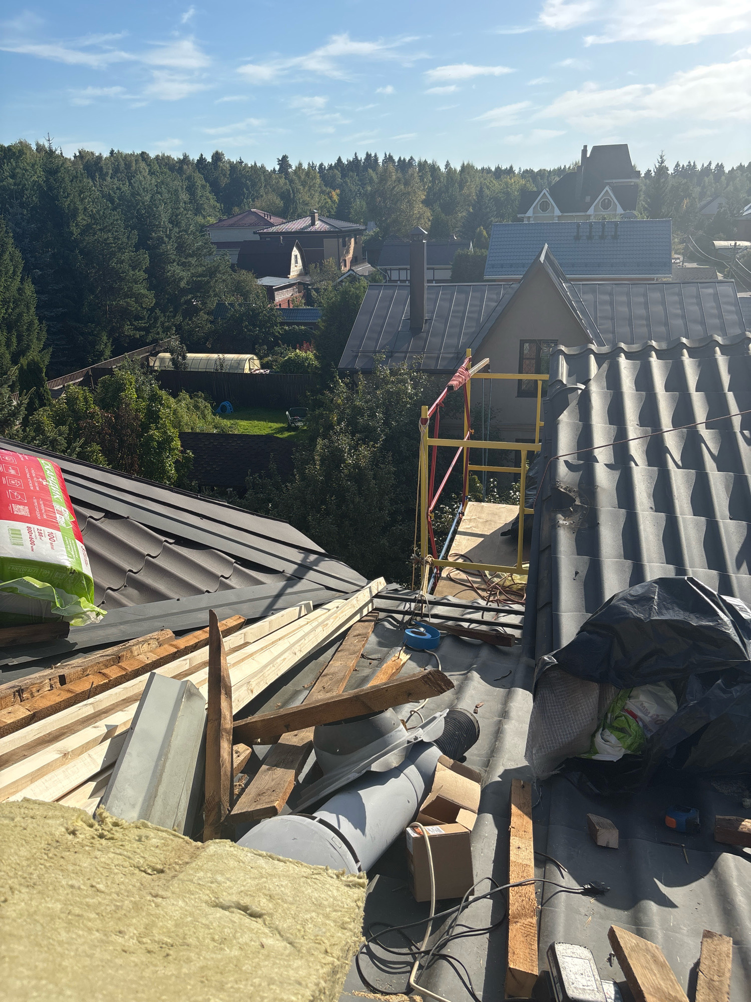 This is a medium-shot, high-angle photograph of a residential rooftop under construction in Zhavoronki, Russia. The scene is dominated by dark gray, wavy roofing tiles and various construction materials. In the foreground, a large roll of light green insulation is visible on the left, partially obscuring the roofing. Scattered across the roof are wooden planks of various sizes, some appearing weathered and others newer. A large gray ventilation pipe is also present, along with several cardboard boxes and black plastic sheeting. A yellow and red scaffolding structure is erected on the right side of the image, with electrical cords and a blue tape measure visible among the debris. In the background, a dense forest of green trees frames several houses with pitched roofs, under a bright blue sky with scattered white clouds. The sun casts distinct shadows across the rooftop. No people are visible in the image.