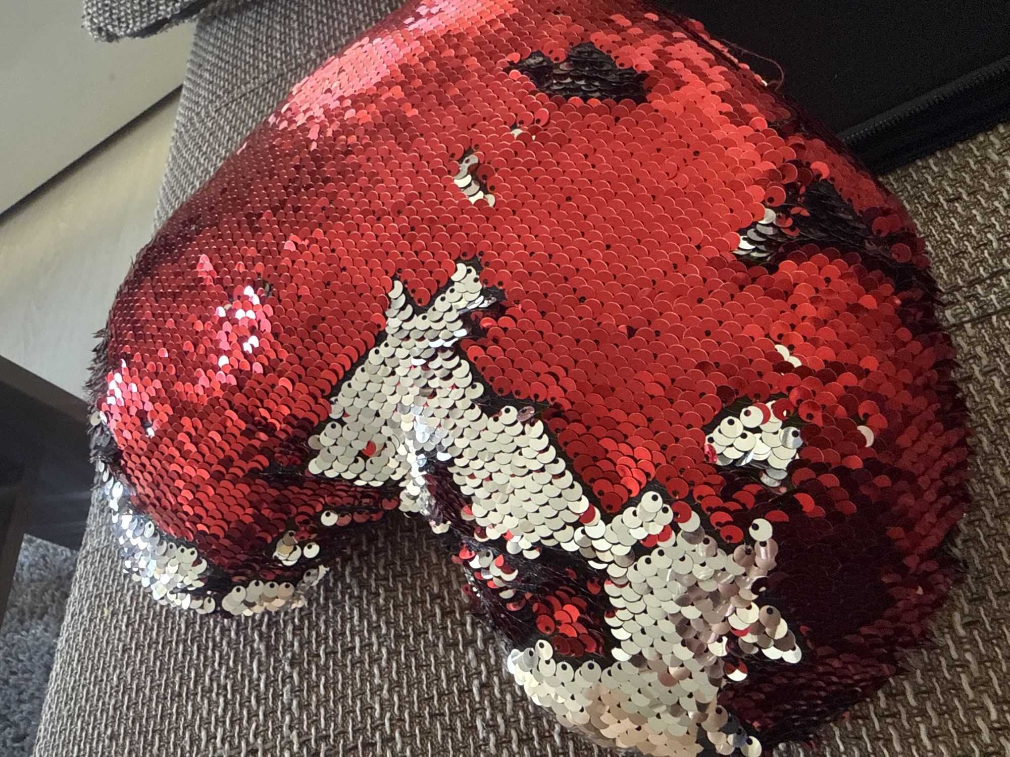 A close-up shot shows a heart-shaped sequin pillow with a design of a cat. The pillow is red on one side and silver on the other, with the cat's face and body appearing as silver sequins against the red background. The pillow is resting on a textured, brown couch. The scene is indoors, and no specific location in Sofia, Bulgaria, is discernible.