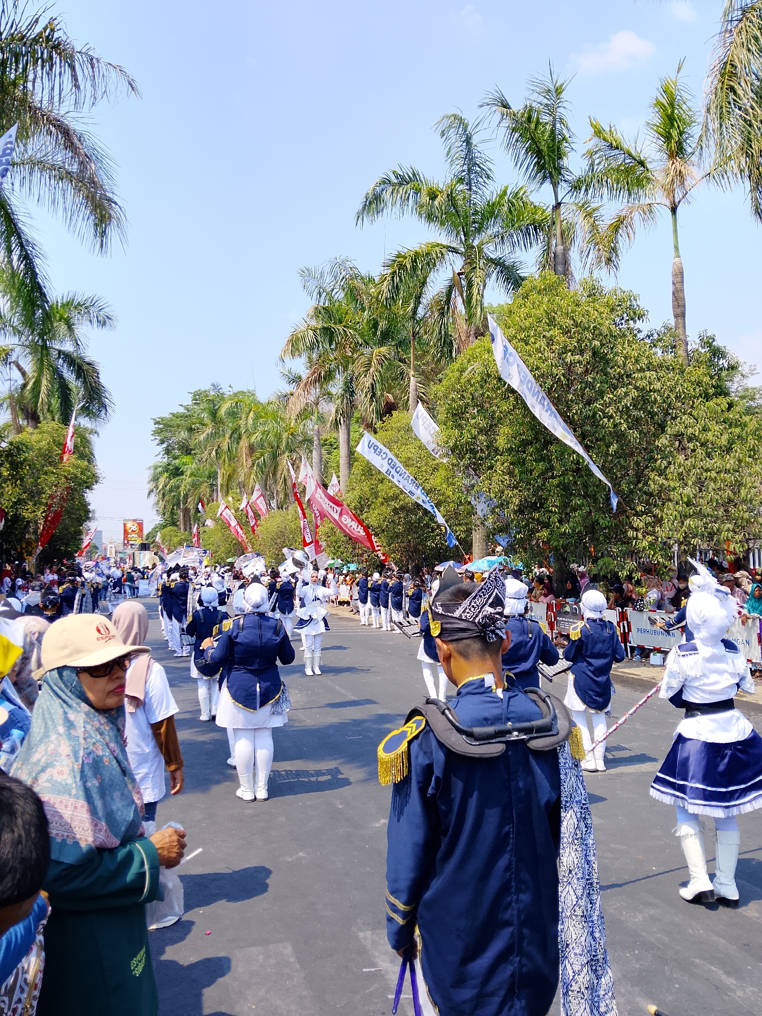 The image depicts a marching band parade in the city of Balun, Indonesia. A group of individuals in matching navy blue and white uniforms are marching down a paved road lined with palm trees and lush green foliage. Some individuals are playing musical instruments, while others are holding batons. The parade appears to be part of a public event, with spectators visible on the sidelines. Several banners and flags are also present, contributing to the festive atmosphere. The sky is clear and blue, indicating a sunny day.