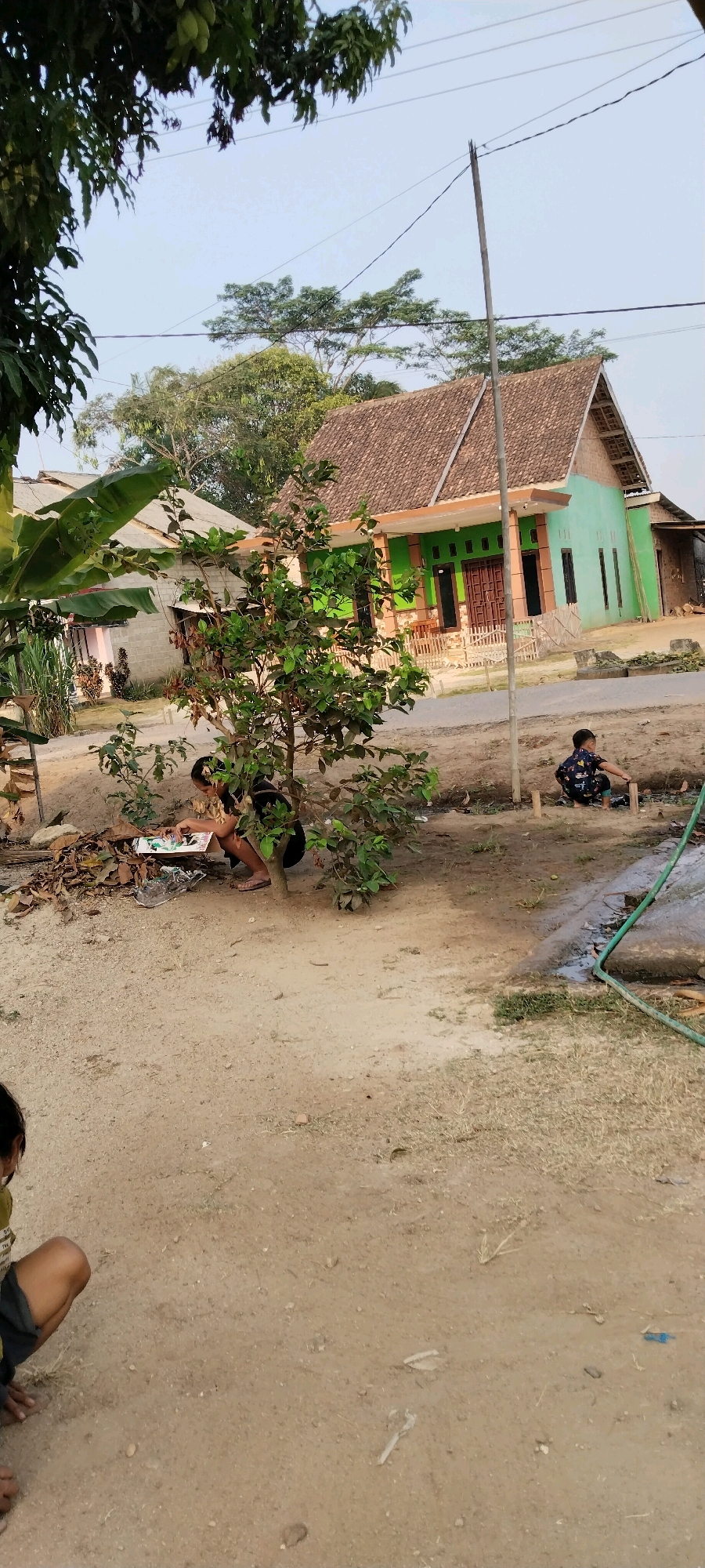 In Tanjung Ratu Ilir, Indonesia, a scene unfolds in a rural setting. Two children are engaged in activities outdoors. A younger child, wearing a dark t-shirt with a colorful pattern and light-colored pants, squats near a wooden structure, seemingly playing with small wooden sticks. Further to the left, a child is crouched near a small tree, looking down at a flat, rectangular object on the ground, possibly a piece of paper or a board. The ground is covered with sand and dry leaves. In the background, a modest green house with a brown tiled roof stands prominently, flanked by other houses. Lush green trees and foliage surround the structures. A utility pole with wires stretches across the frame, and a green hose is visible on the right side of the image. The overall environment appears to be a residential area with a natural, undeveloped feel.