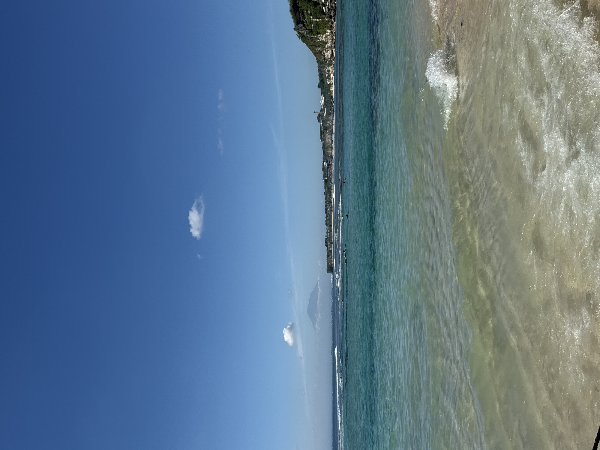 The image depicts a beach scene in Kuta Selatan, Indonesia. The sky is clear blue with scattered clouds. Along the shoreline, the ocean transitions from a clear turquoise to a foamy white as the waves break on the sand. Buildings can be seen along a cliff that borders the beach. Some people are visible in the water.