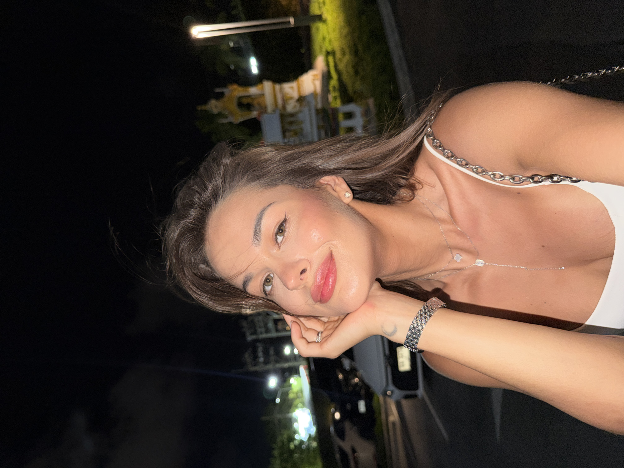 A woman poses outdoors at night in Choeng Thale, Thailand. She rests her face on her hand, which displays a silver bracelet and a ring. She wears a white top with a silver chain strap and a pendant necklace. In the background, trees, streetlights, and parked vehicles are visible, indicating an urban environment. A golden Thai spirit house is partially visible.