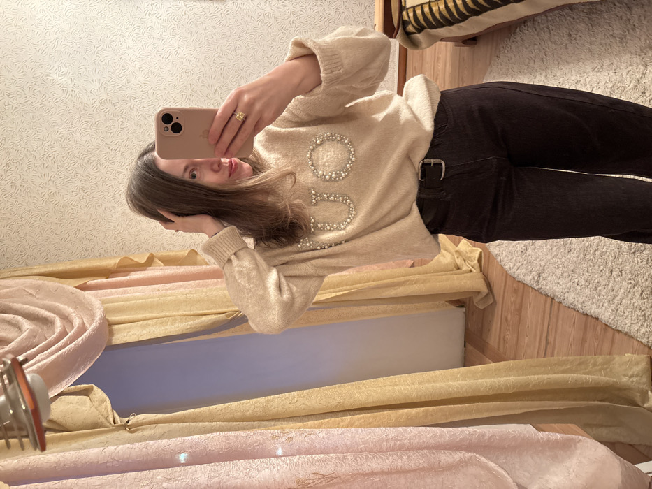 A mirror selfie captures a young woman standing indoors, holding a light-colored smartphone to take her picture. The image is oriented sideways, with the woman's head on the left and her feet extending to the right.
The woman has long, straight light brown hair and a neutral expression, looking directly into the phone's camera reflection. Her face is partially obscured by the phone. She is wearing a soft, light beige or cream-colored knitted sweater with ribbed cuffs. Prominently displayed on the front of the sweater are large, decorative letters formed by white pearls or beads. The letters "GU" are clearly visible, followed by what appears to be a "C" or "O," suggesting a brand name like "GUCCI." She is also wearing dark brown or black trousers, secured with a brown belt featuring a silver-toned buckle. A golden ring is visible on the ring finger of her right hand, which is holding the phone.
The background behind her offers glimpses of various indoor elements. To her left (top of the image when viewed sideways), there's a light-colored wall covered in textured wallpaper with a subtle, swirling or leafy pattern. To her right (bottom of the image when viewed sideways), the floor consists of light wooden planks, partially covered by a fluffy, off-white or cream-colored shaggy rug.
On the far left side of the image (bottom when viewed sideways), there are multiple layers of curtains. The topmost layer visible is a gold or yellow crushed fabric, beneath which is another layer of pinkish, possibly velvet or crushed fabric. A gap between the curtains reveals a light blue or lavender wall or surface. In the bottom left corner, a metallic object with a circular design and a reddish accent is visible, possibly part of a light fixture or decor. In the upper right corner (far right when viewed sideways), part of a piece of furniture, possibly a bed or bench, is visible, draped with a striped blanket in shades of brown and beige. The lighting appears to be ambient indoor light.