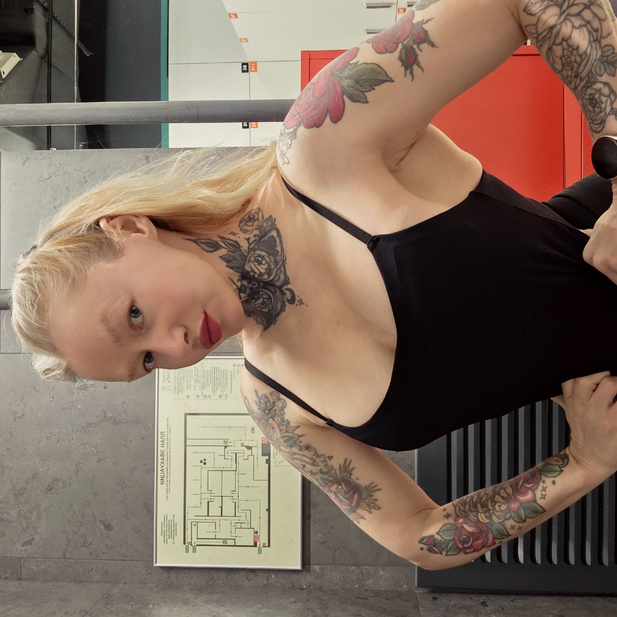 A woman with blonde hair and multiple tattoos poses indoors in Saint Petersburg, Russia. She is wearing a black tank top. A framed diagram with Cyrillic text is visible on the wall, indicating an evacuation plan. Lockers are in the background, with a red section visible. The woman appears to be holding onto a horizontal bar.