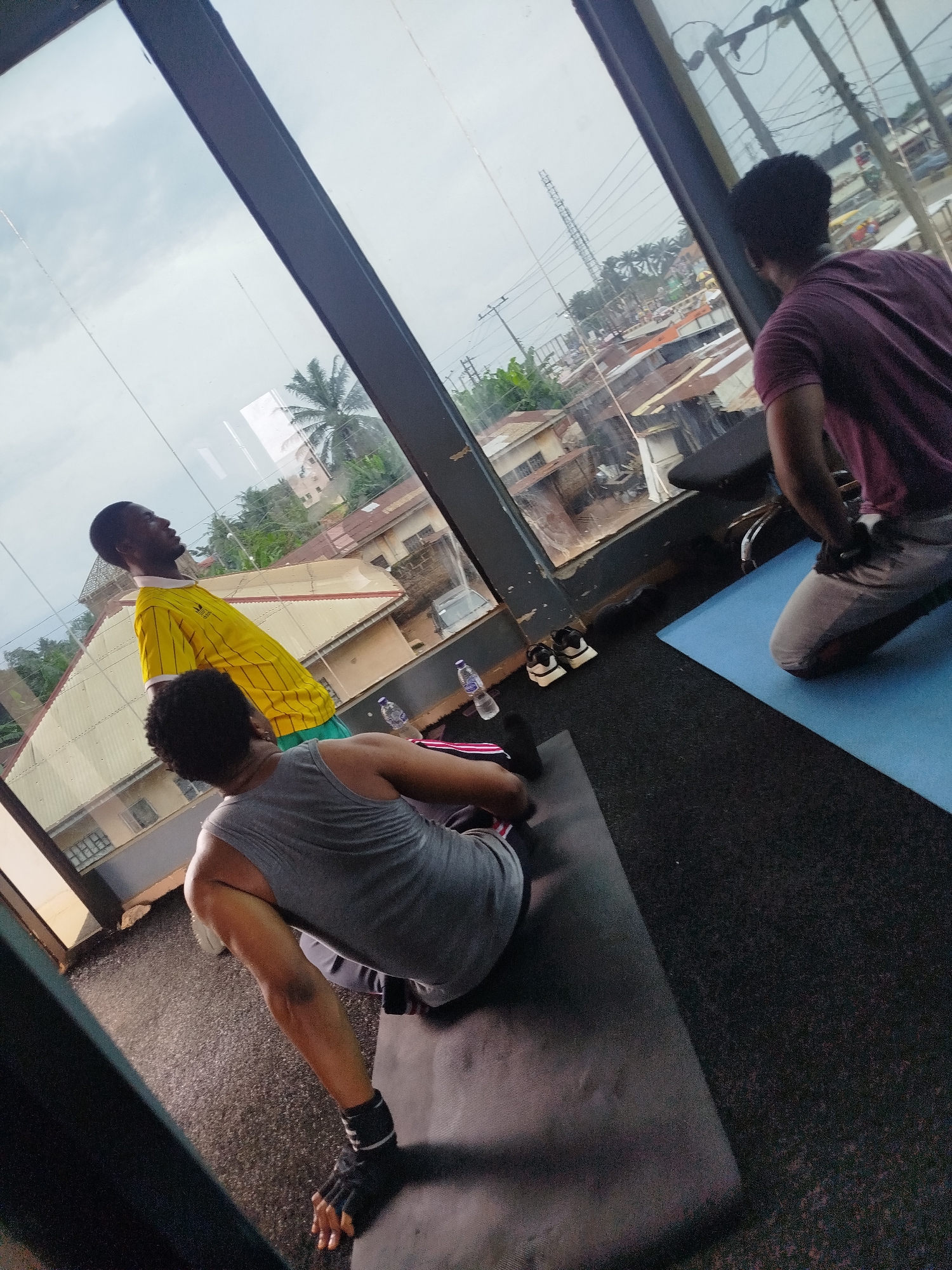 In Uselu, Nigeria, three individuals are engaged in a fitness activity near a large window offering a view of an urban landscape. Two people are on yoga mats on the floor. One person, wearing a gray tank top and fingerless gloves on their hands, is in a seated position on a black yoga mat, facing away from the camera. Their legs are bent and crossed at the ankles, and they appear to be stretching or resting. Another person, wearing a purple t-shirt and light-colored pants, is kneeling on a blue yoga mat, facing the window and looking out at the city. A third person, wearing a yellow and white striped t-shirt and green shorts, stands between the other two, looking upwards and away from the camera. Water bottles and a pair of athletic shoes are visible on the floor near the window. The environment suggests an indoor gym or fitness space with large windows overlooking a densely built urban area with corrugated metal roofs and some vegetation. The sky is overcast.