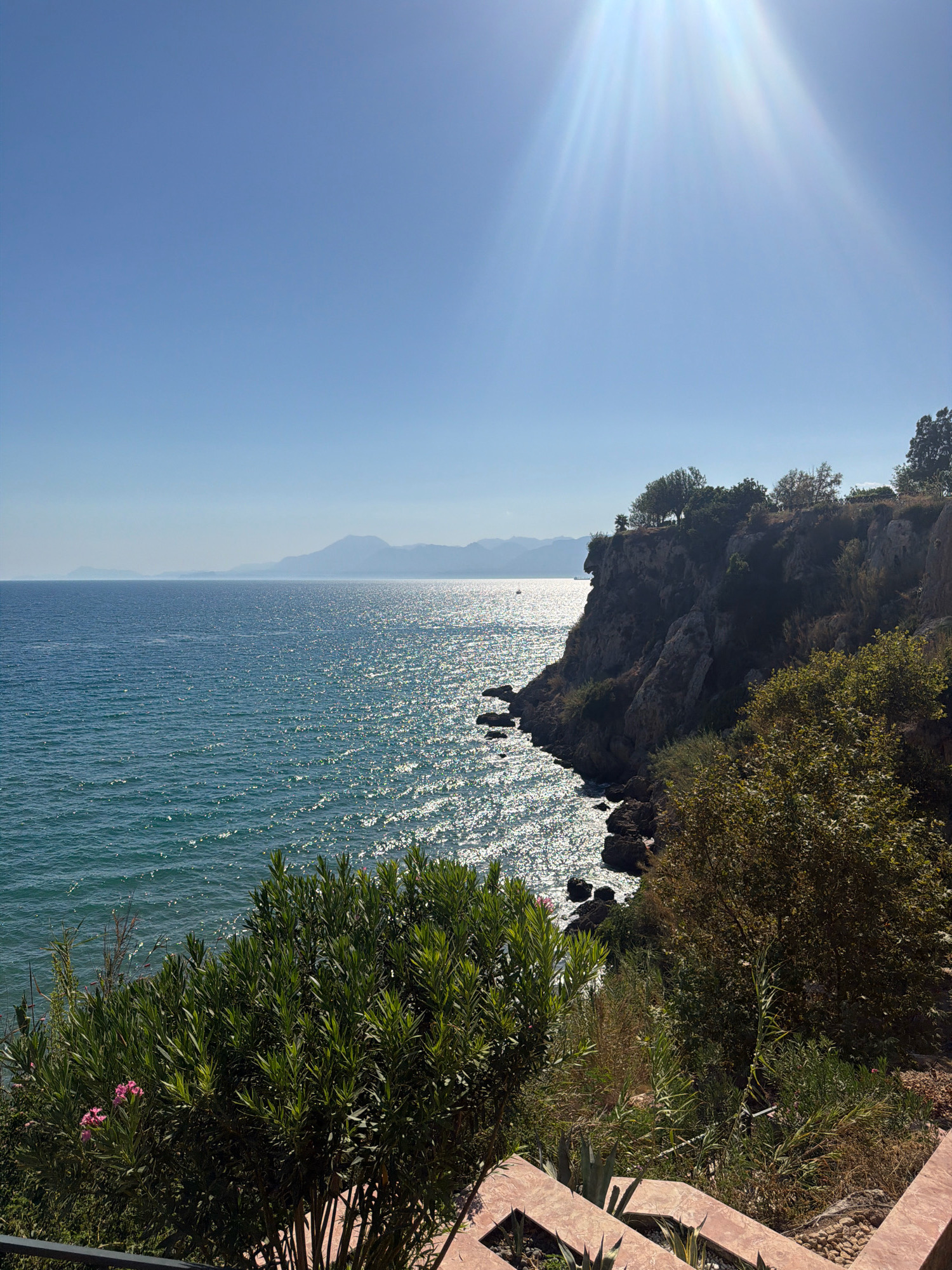 The image captures a scenic coastal view in Antalya, Turkey, likely from a high vantage point overlooking the Mediterranean Sea. The foreground features lush green foliage, including an oleander bush with pink flowers, and a stone staircase. Dominating the mid-ground is a dramatic cliff face dotted with trees and shrubbery, dropping steeply to the sea below. The sea itself is a deep blue, shimmering with sunlight, with small waves lapping against rocky outcrops at the base of the cliff. In the distance, a hazy mountain range stretches across the horizon under a clear, bright blue sky. The sun is high, casting bright rays that create a strong glare on the water. No people or animals are visible in the scene.