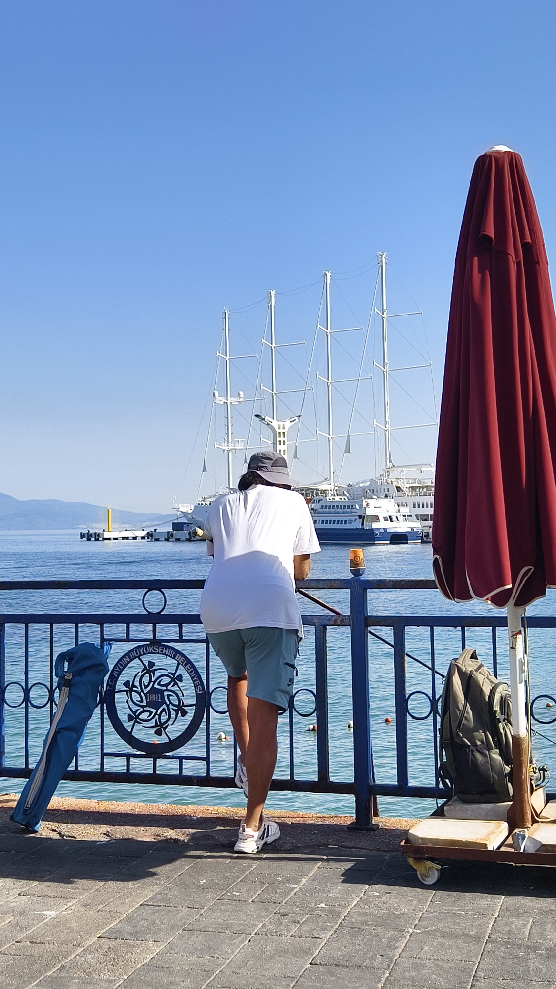 The image depicts a person wearing a white t-shirt, light blue shorts, a hat, and white sneakers, leaning on a blue railing. The person is facing away from the camera, looking out at a body of water. In the background, a large sailing ship with multiple masts is visible, along with other boats and a distant shoreline. To the right, a large, folded red umbrella is prominently featured. A blue bag is hanging on the railing to the left, and a green backpack is placed on a trolley to the right. The ground is paved with gray stones. The scene appears to be a waterfront promenade in Kuşadası, Turkey, likely near the marina or a public viewing area given the presence of boats and leisure-oriented amenities like the umbrella. The sky is clear and blue.