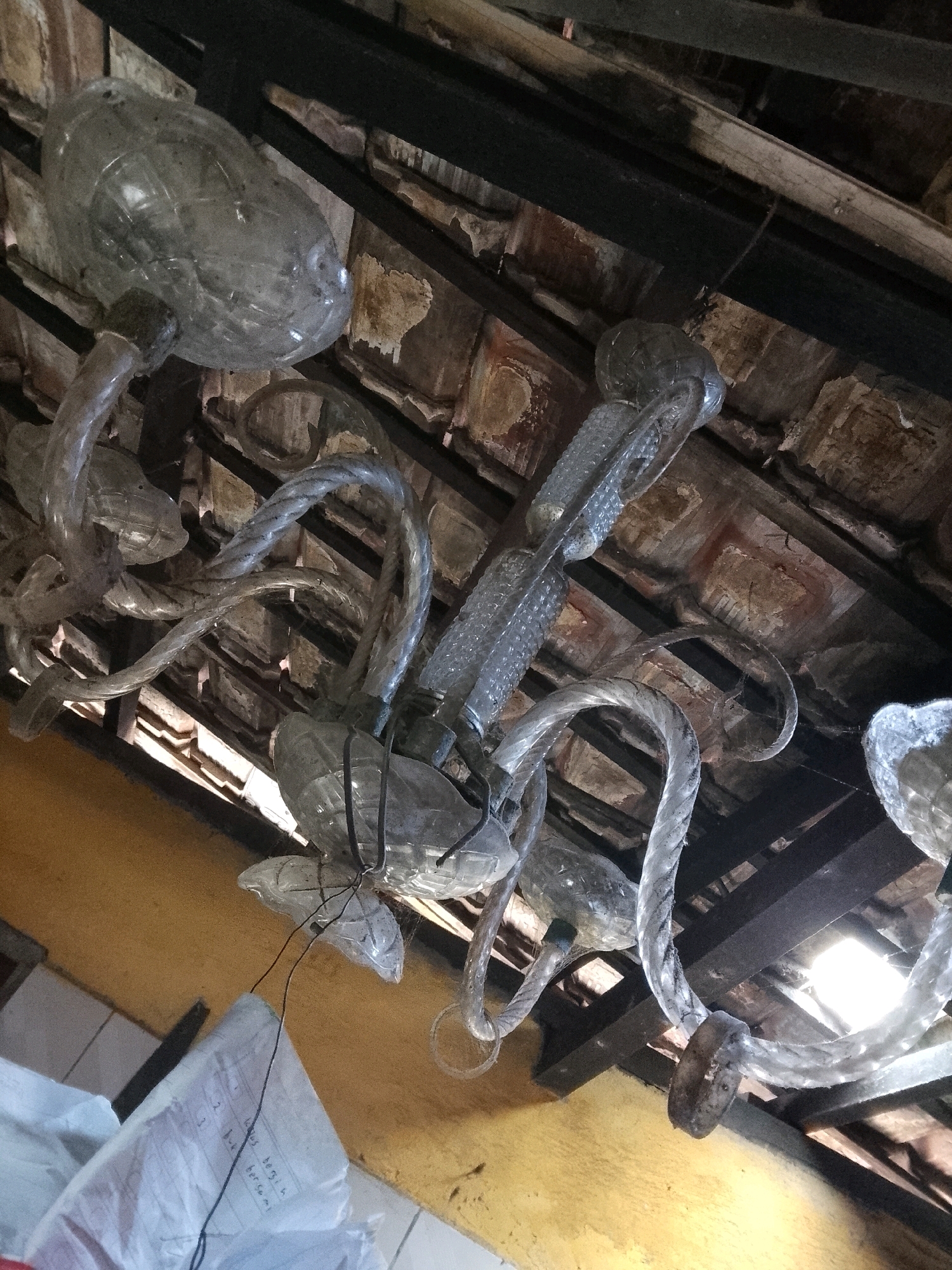 The image depicts a rustic chandelier constructed from repurposed materials, likely plastic bottles and possibly glass tubes, suspended from a weathered wooden ceiling structure. The chandelier features multiple arms that twist and curve, with what appear to be large, translucent bottle caps serving as the main light-diffusing elements. The ceiling itself is composed of worn wooden beams and exposed terracotta tiles, suggesting an older, possibly outdoor or semi-outdoor structure. Sunlight filters through gaps in the ceiling, creating highlights and shadows. A roll of white paper with handwritten text is visible in the lower left quadrant, attached to a wire that seems to be part of the chandelier's suspension. The overall impression is one of resourceful recycling and unconventional design within an aged architectural setting. This scene is located in Surabaya, Indonesia.