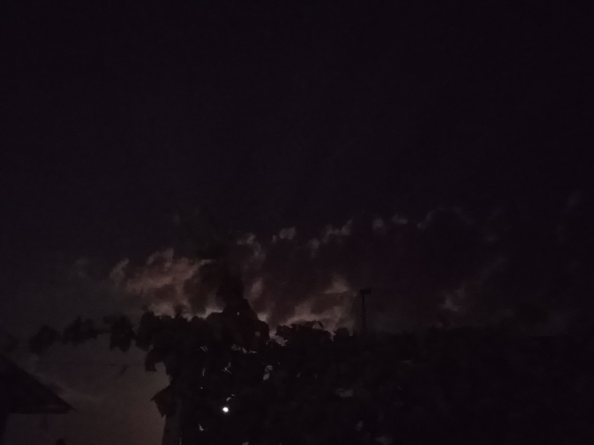 This is a nighttime image, likely taken outdoors, with very dark conditions. The sky is predominantly black with some discernible clouds that are faintly illuminated, possibly by moonlight or distant artificial light. In the foreground, silhouetted against the slightly lighter sky, are dark shapes of trees and foliage. A portion of a structure, possibly a roof or building edge, is visible on the far left of the image. A faint light source, appearing as a small white dot, is present in the lower portion of the frame, near the center. The scene lacks clear details of people or specific urban features due to the low light conditions. It is not possible to determine the exact location within Ucrainca, Moldova, based solely on this image.