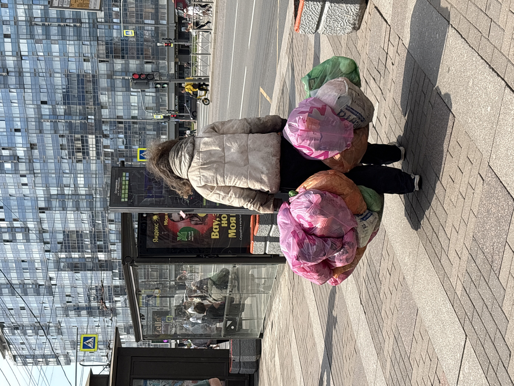 In Saint Petersburg, Russia, on a sunny day, a woman with brown, curly hair, wearing a light-colored quilted jacket and dark pants, walks away from the camera. She is carrying a large bundle of plastic bags filled with items, held in both arms. The bags are brightly colored, predominantly pink, orange, and green, and appear to be full and heavy. The woman is walking on a paved sidewalk made of interlocking stones. In the background, a modern glass-fronted building with many windows dominates the skyline. A bus stop shelter with advertisements, including one for "Yandex," is visible to the left of the woman. Traffic lights and street signs, including a pedestrian crossing sign, are also present on the street behind her. People can be seen in the background near the bus stop and walking on the sidewalk across the street. The overall scene depicts an urban environment with a person engaged in carrying goods.