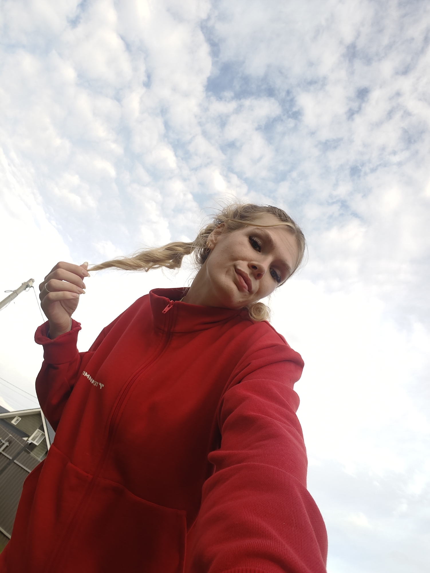 The image shows a young woman with blonde hair styled in two braids, wearing a red zip-up hoodie. She is holding one of her braids with her left hand, and her right hand is out of frame. The woman is looking towards the camera with a slight smile and her head is tilted. She is outdoors, with a cloudy sky in the background. A power pole and a building are partially visible on the left side of the image. The perspective is from a low angle, looking up at the woman and the sky. The overall scene suggests a casual, outdoor setting.
