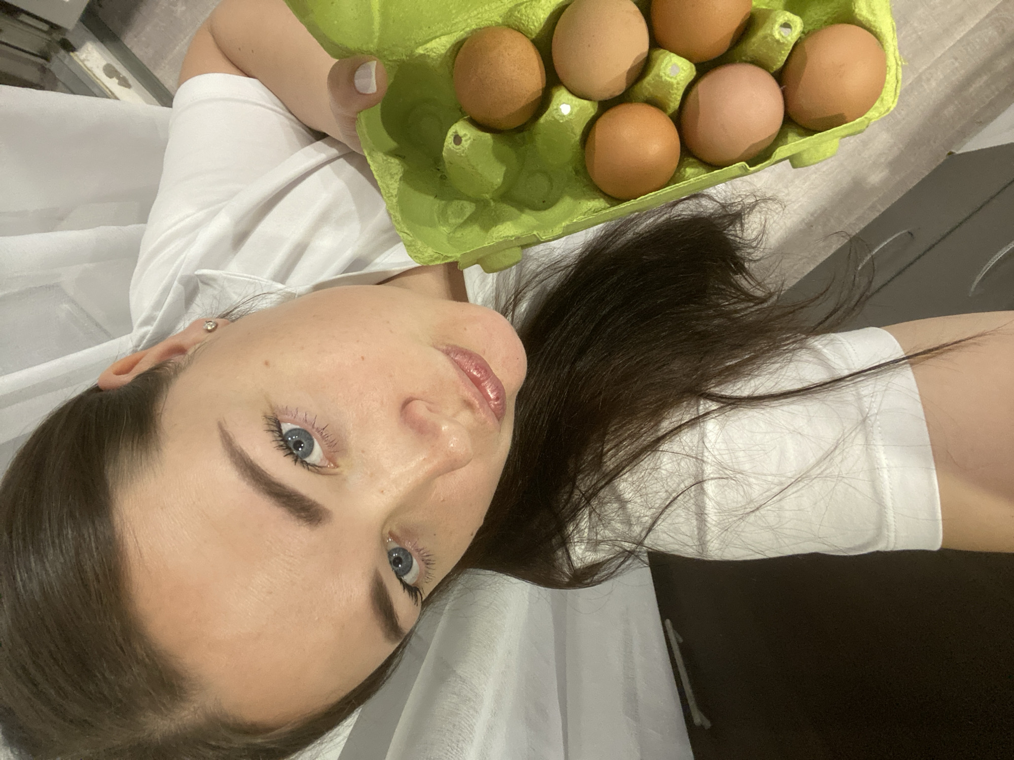 The image shows a woman holding a green egg carton with six brown eggs. The woman has fair skin, blue eyes, and dark hair. She is wearing a white t-shirt. The background includes what appears to be a table surface with some grey and brown tones. There is no evidence of Sochi, Russia.