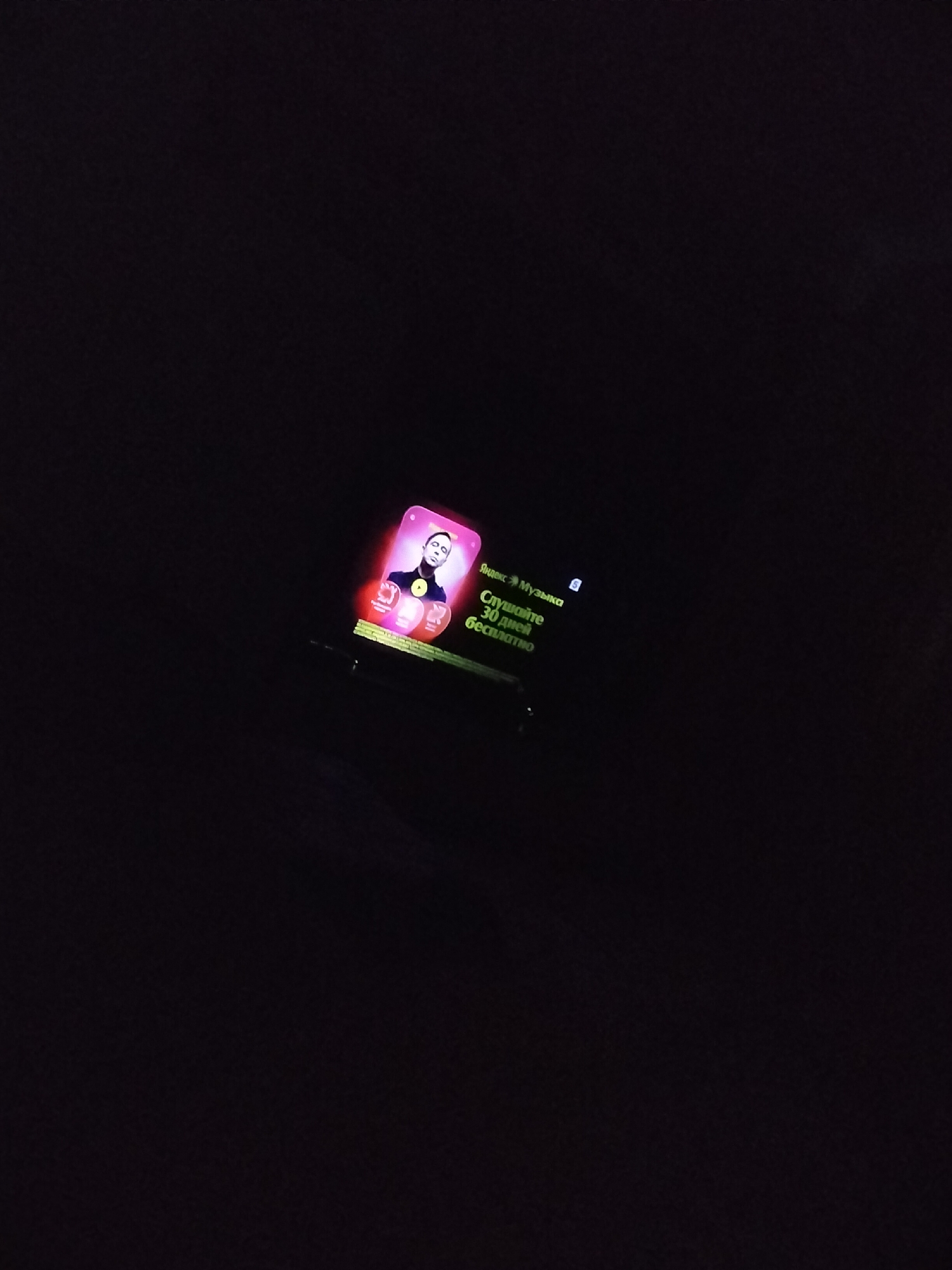 A smart watch displays a pink screen with an image of a person and text. The text is in Russian and reads "Яндекс Музыка" and "Слушайте 30 дней бесплатно". The watch is on a dark surface, likely a wrist, in a dimly lit environment. The scene appears to be indoors, possibly in a residential setting in Nizhnedonskoy, Russia, though this specific location cannot be definitively confirmed from the image alone.