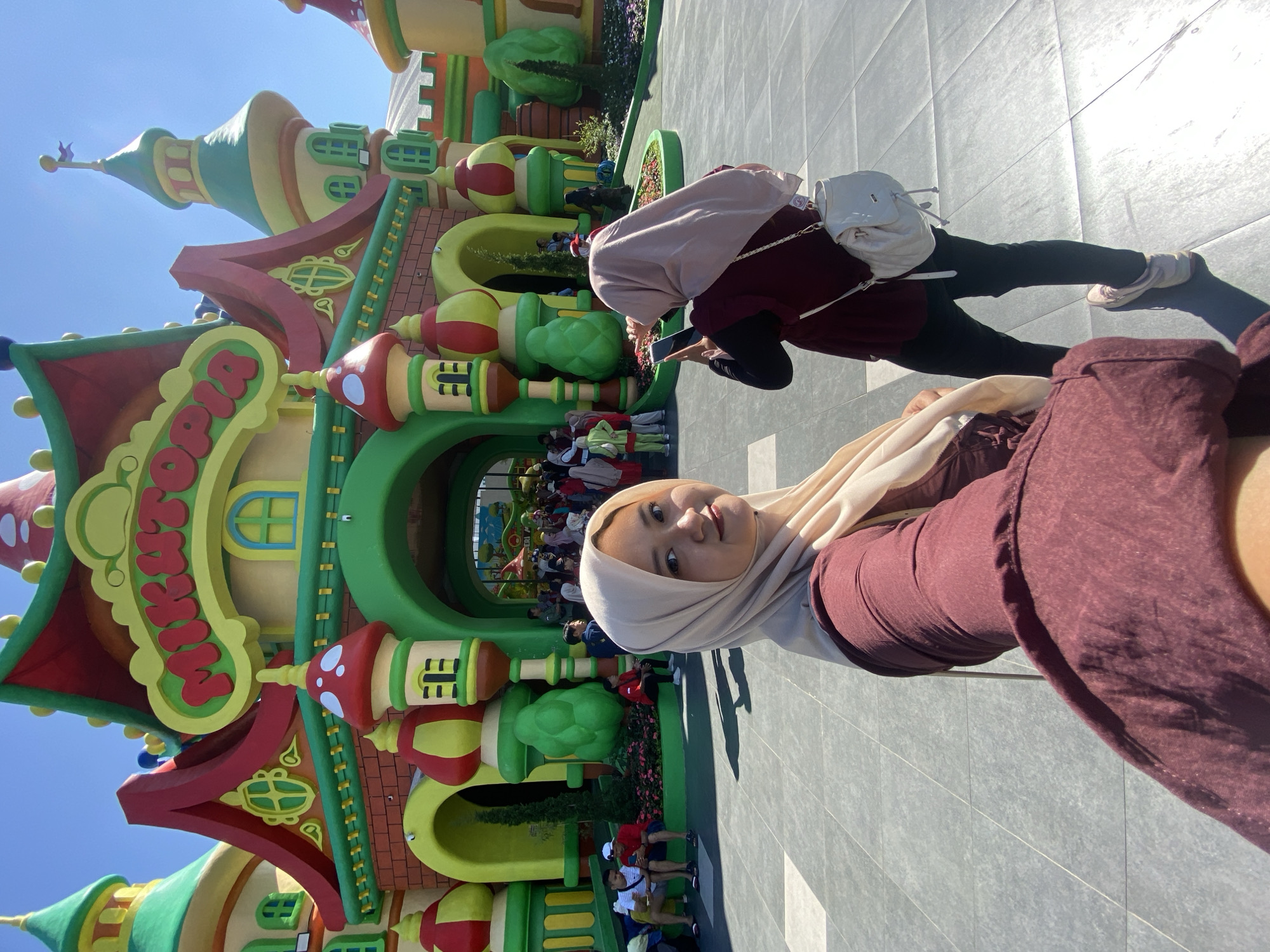 This image captures a bright daylight scene at the "Mikupia" attraction within Jatim Park 3, located in the city of Batu, East Java, Indonesia.
In the foreground, a woman wearing a maroon top and a light beige hijab is taking a selfie, holding her phone with her right arm extended towards the camera. She looks directly into the lens with a slight smile. To her right and slightly behind, another woman with a light pink hijab and a maroon top, black pants, and light-colored sneakers is visible from the back, holding a black smartphone to take a photo of the colorful building. She carries a small white backpack on her left shoulder.
The background is dominated by the vibrant, whimsical architecture of the "Mikupia" building, which features multiple turrets and arched entrances. The structure is painted in bright primary colors, predominantly yellow walls, green roofs, and red accents. The name "MIKUTOPIA" is prominently displayed in red letters on a yellow banner across the central facade. Mushroom-shaped decorations in red and white are visible on some of the building's turrets. Further back, numerous individuals are gathered near the building's entrances, some sitting on ledges, others walking or standing, indicating a public leisure setting. Small landscaped areas with green bushes and flowers are present along the base of the building. The ground is paved with alternating light and dark gray square tiles. The sky above is clear and bright blue. Strong sunlight casts distinct shadows on the ground.