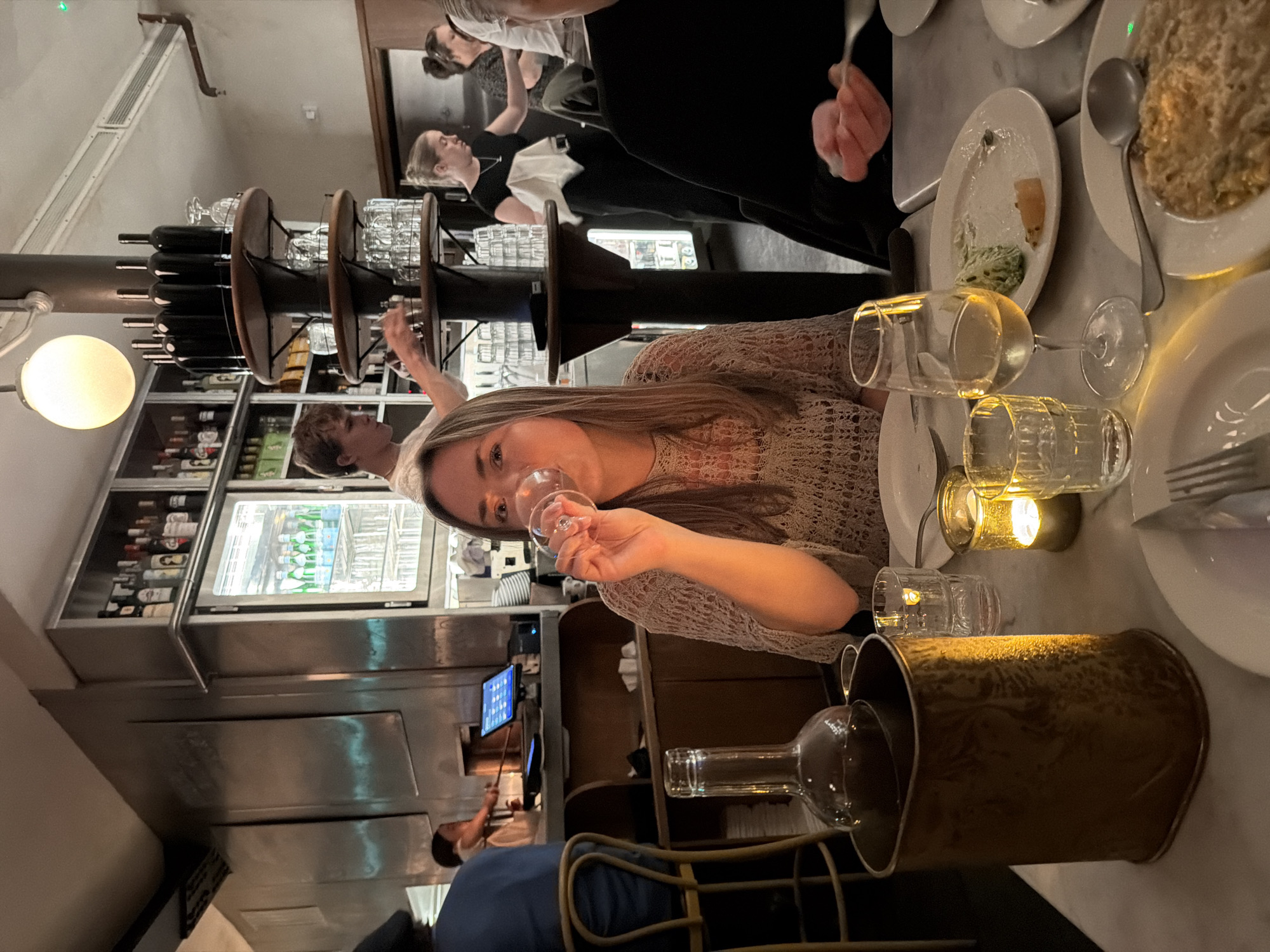 A dimly lit interior scene shows a group of people dining at a restaurant. The restaurant is characterized by its modern decor, featuring dark wooden elements, glass wine racks, and illuminated beverage displays. A woman is sitting at the table, holding a wine glass, and partially facing the camera. Other diners are present, including a person wearing a black shirt and another in a blue shirt seated in the background. The table is set with plates, silverware, and several wine glasses, as well as a metallic bucket that holds an empty wine bottle. Based on the aesthetic of the scene, and architectural details, the restaurant is likely located in London, United Kingdom.