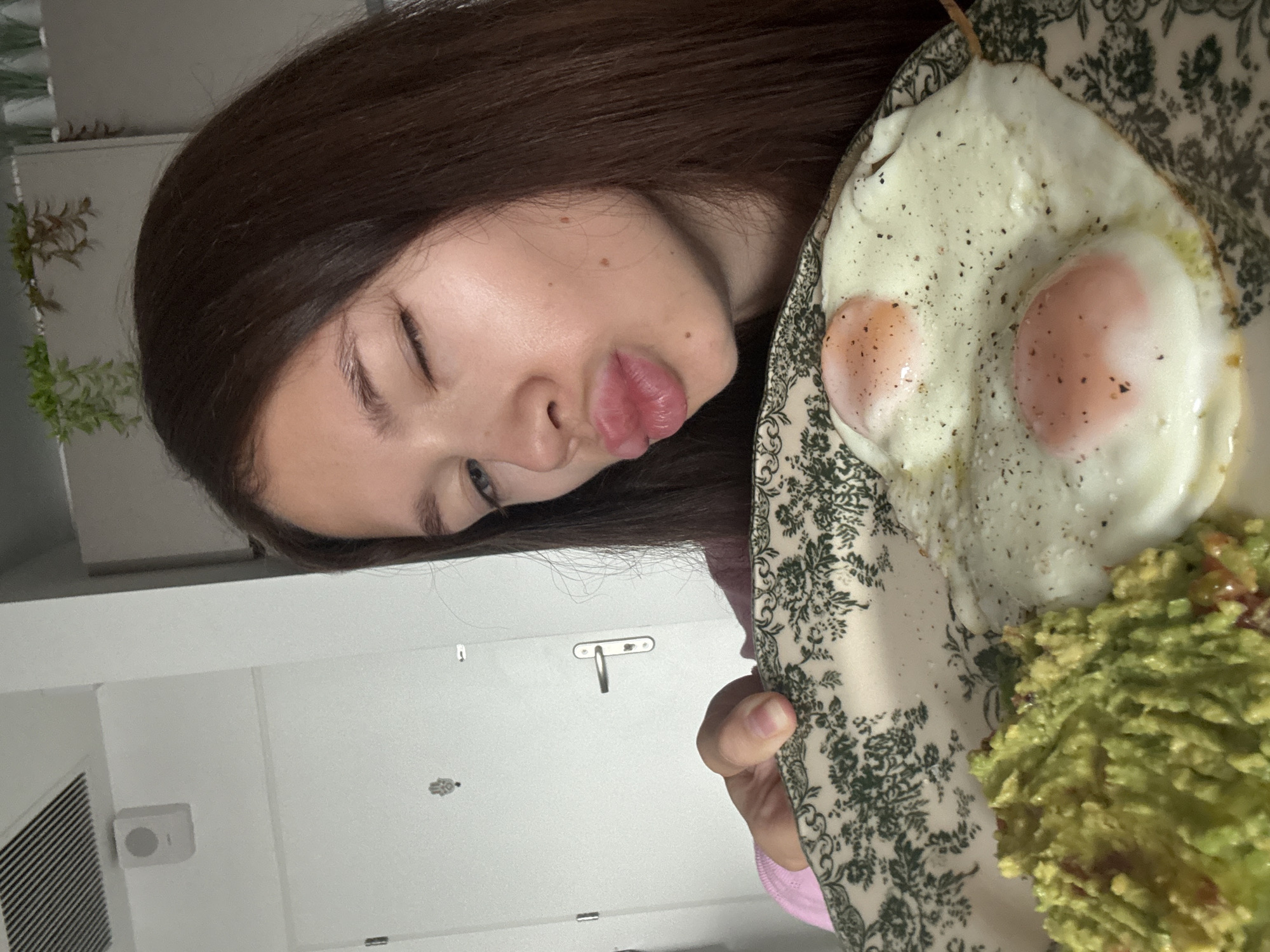 A young woman with long brown hair holds a plate of food. On the plate are two fried eggs sprinkled with pepper, and a mound of guacamole. The woman is making a kissing face towards the food. The background appears to be a white room with a vent and a cabinet. It is not possible to verify the location is Ramat Gan, Israel, from the given image.