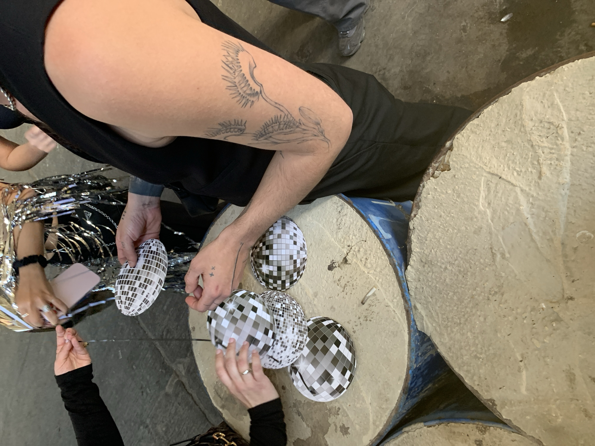 The image captures a close-up, high-angle view of multiple individuals gathered around a makeshift table in an indoor or semi-enclosed industrial-style setting. The specific location within Tel-Aviv, Israel, is not discernible from the provided visual information. The table is constructed from several blue industrial barrels topped with a textured, off-white concrete-like surface, upon which a discarded cigarette butt is visible.
In the foreground, an individual's right arm, clad in a black sleeveless garment, displays a prominent tattoo depicting multiple Venus flytraps. This individual's hands, marked with small 'x' and a horizontal line tattoo on the right hand, hold an elongated, oval-shaped object patterned like a disco ball. The person wears black trousers, and a grey athletic shoe is partially visible at the bottom right.
To the mid-left, another person's hands, wearing a black long-sleeved top and a ring on their left ring finger, are positioned around three spherical, disco-ball-patterned objects resting on the table, appearing to arrange them. In the background center, a third individual, partially visible and adorned in a shimmering silver, fringe-like garment, is seen holding a small pink object and a thin dark string. The floor is a dark, possibly concrete, surface.