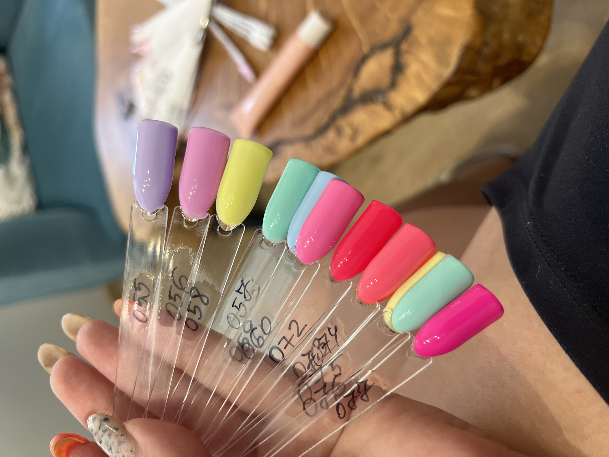 The image shows a close-up of a person's hand holding a fan of nail polish samples. The hand has polished nails, some with a speckled design and others with bright orange. The nail polish samples are on clear plastic sticks, each with a handwritten number. The colors visible are purple, pink, yellow, turquoise, light blue, bright pink, and neon red. In the background, out of focus, is a wooden table, a light-colored tube, and some manicure tools. The scene is likely within a nail salon.