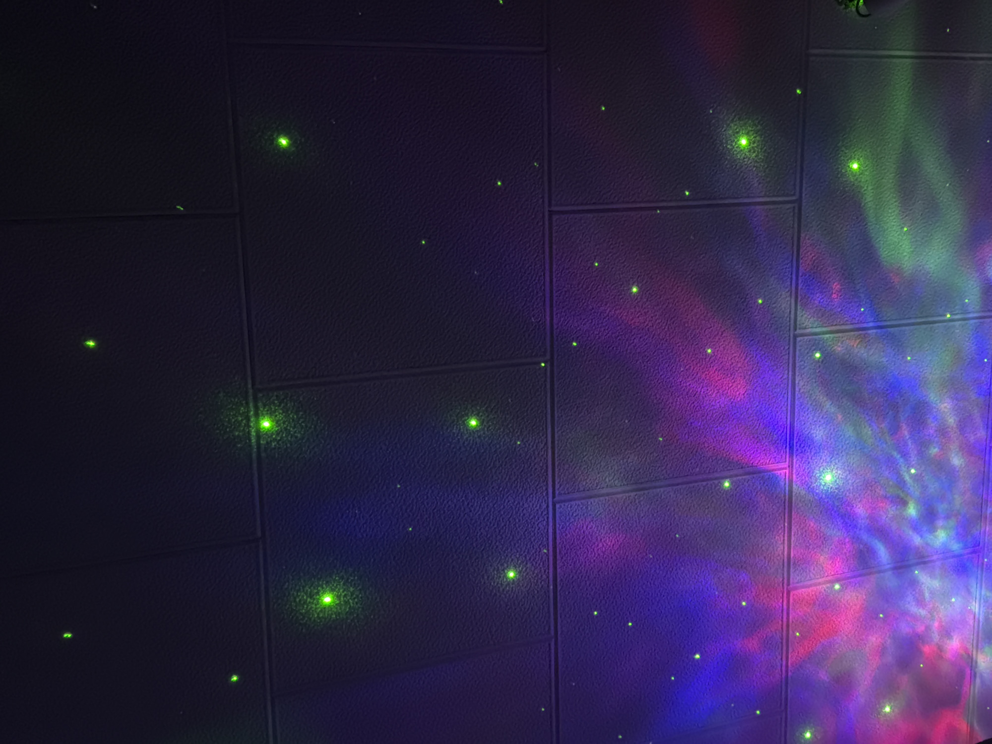 The image shows a section of a wall covered in square tiles. The tiles are dark in color, possibly dark gray or black, and have a textured surface. A pattern of small, bright green dots is scattered across the surface of the tiles. On the right side of the image, there is a colorful, nebulous light projection with hues of blue, purple, red, and green. The light appears to be illuminating the tiles. The wall appears to be indoors. There is no information to suggest that this image was taken in Pazardzhik, Bulgaria.