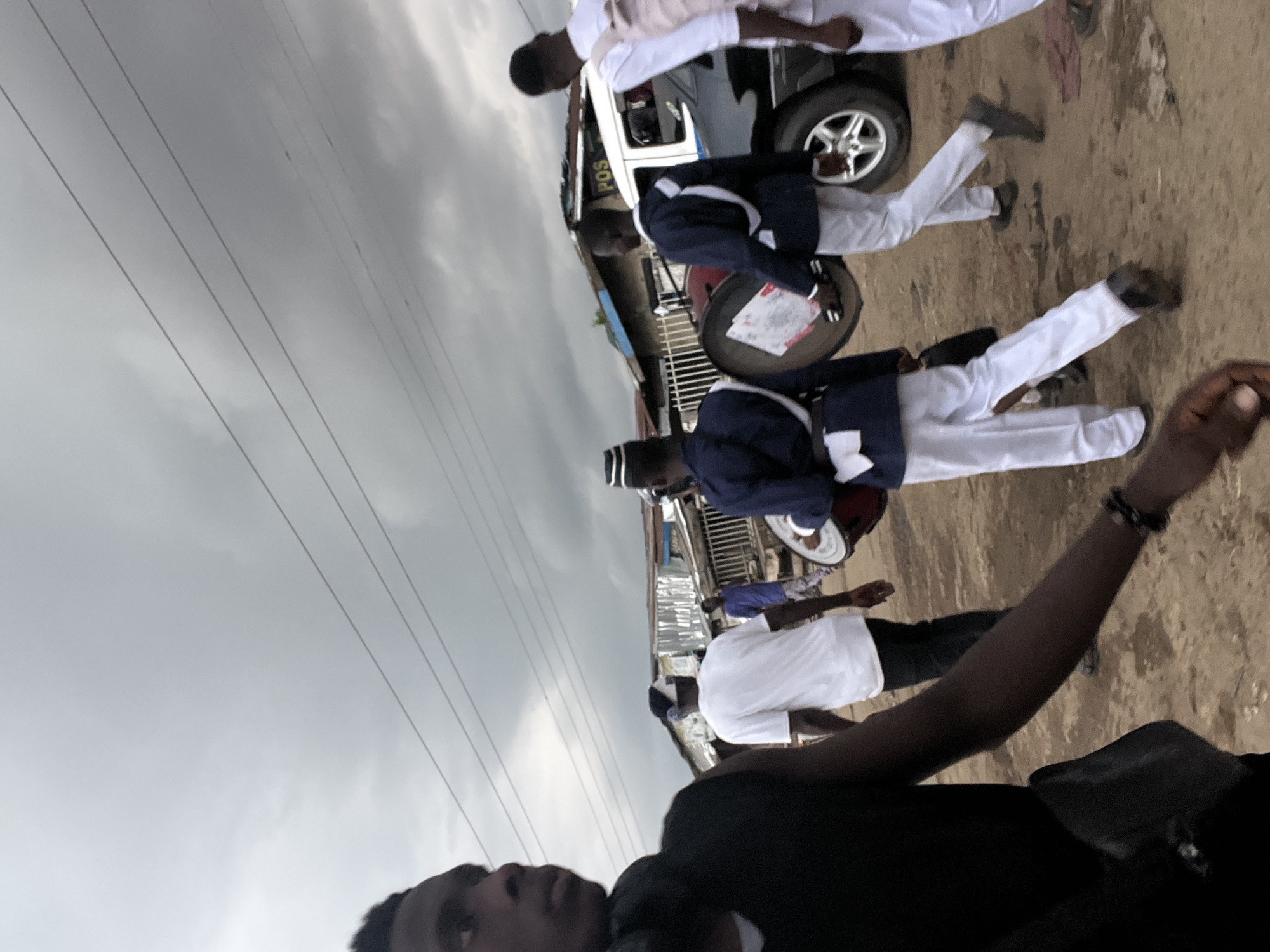 A medium-wide shot captures a group of people walking down a dirt road in a dusty, urban environment. The sky is heavily overcast, with dark, grey clouds suggesting impending rain. In the foreground, a young Black man is partially visible on the left, wearing a black sleeveless shirt and a black crossbody bag. He is looking to his left, with his left arm extended forward. Behind him, a person in a white t-shirt and dark pants is walking away from the camera. Further back, several individuals are dressed in what appears to be a marching band uniform, consisting of navy blue jackets with white sashes and white pants. Two individuals are carrying drums. One drum is large and cylindrical, leaning against the shoulder of a person in a blue jacket. Another drum is visible in front of this person. A white vehicle, possibly a van, is parked on the right side of the road, with "POS" visible on its side. The background consists of corrugated metal structures and some fencing, indicative of a commercial or residential area. The overall scene suggests a community event or procession in Asaba, Nigeria, under a dramatic sky.