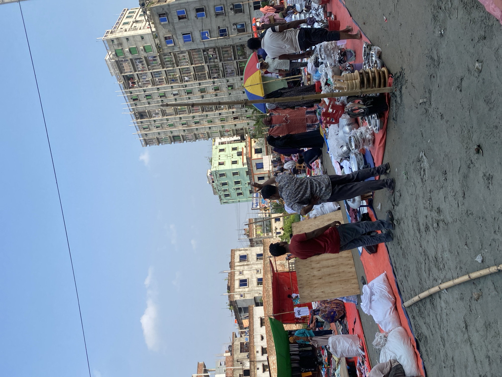 The image depicts an outdoor market scene in Dhaka, Bangladesh. Rows of vendors display goods on red tarps laid out on the ground. Merchandise includes clothing, kitchenware, and other household items. People are browsing the stalls, and a few are engaged in conversation, possibly bargaining. Buildings are visible in the background, varying in height and construction, from low-rise brick structures to taller, multi-story concrete buildings. The sky is mostly clear with a few clouds. The location appears to be a street or open area within a densely populated part of Dhaka.